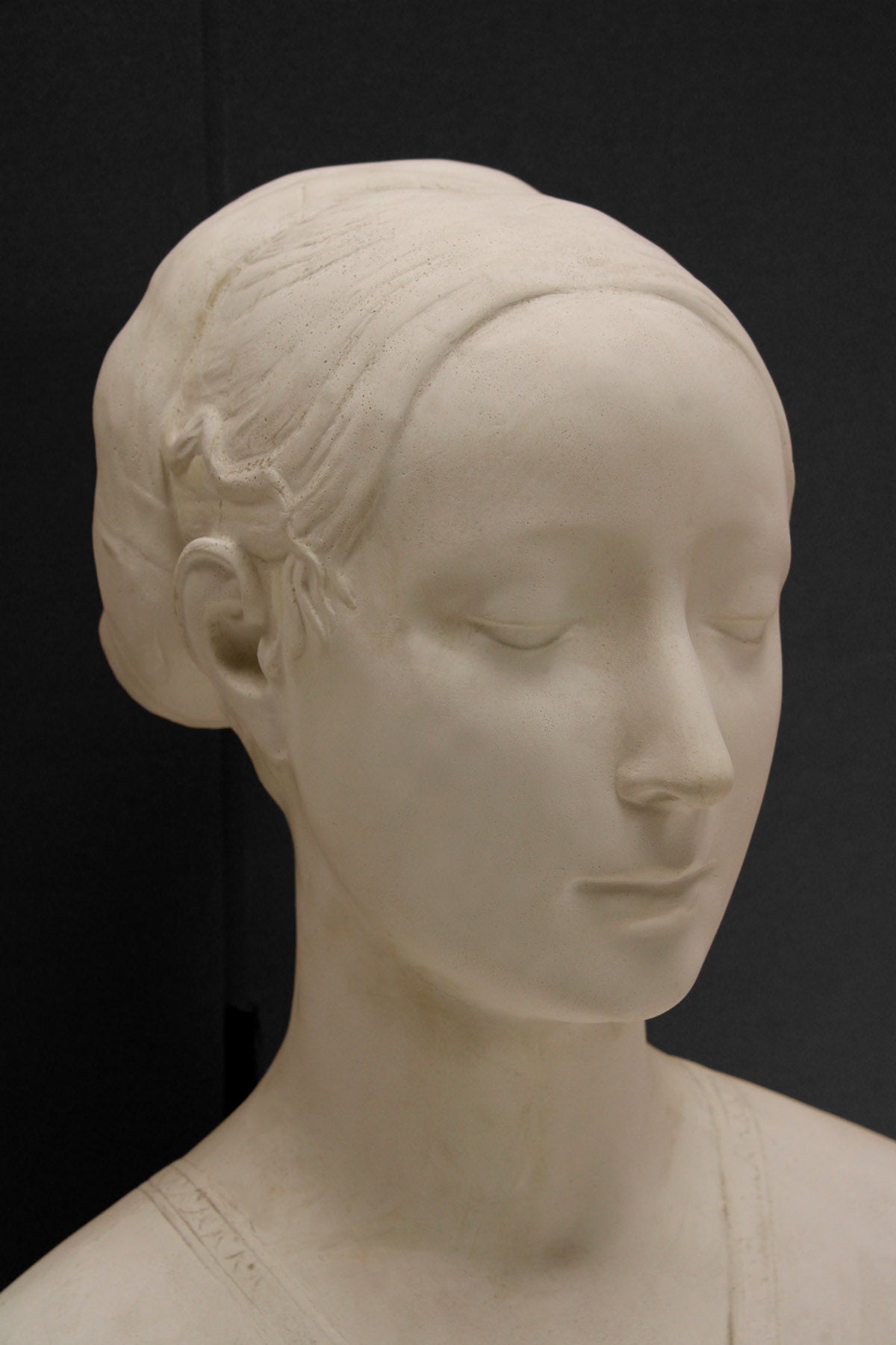detail photo of white plaster cast sculpture of female bust cropped at shoulders with decorative base against black background