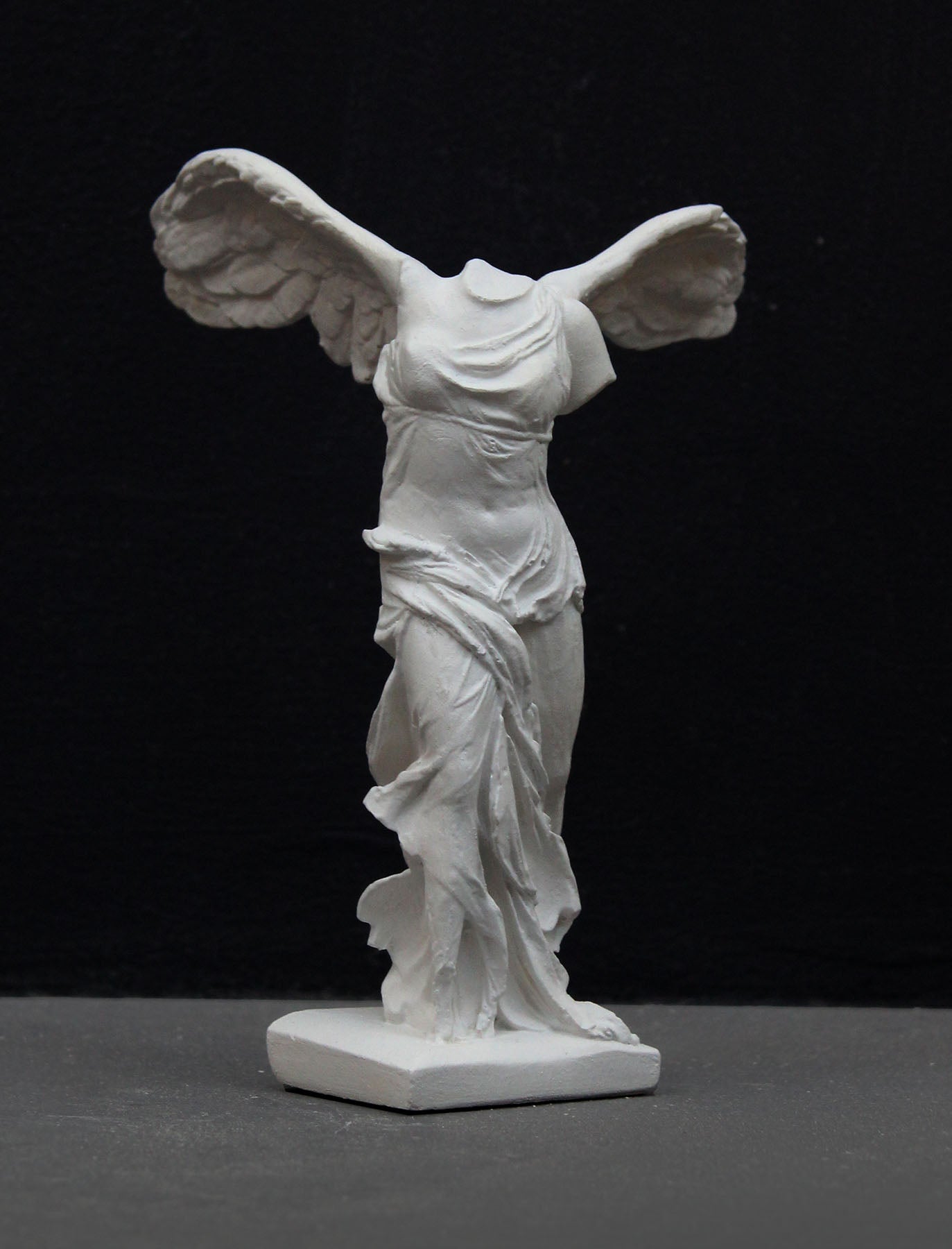 photo of white plaster cast sculpture of winged, headless female figure with flowing drapery on a gray background