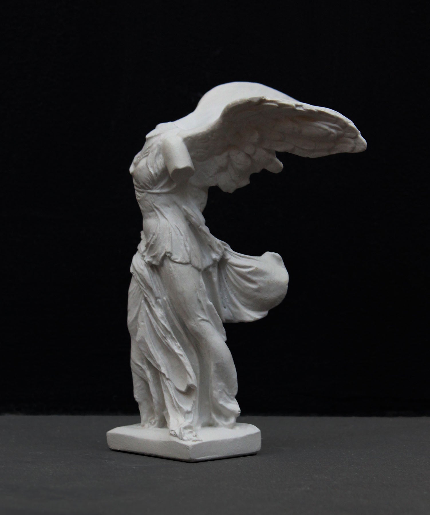 photo of white plaster cast sculpture of winged, headless female figure with flowing drapery on a gray background