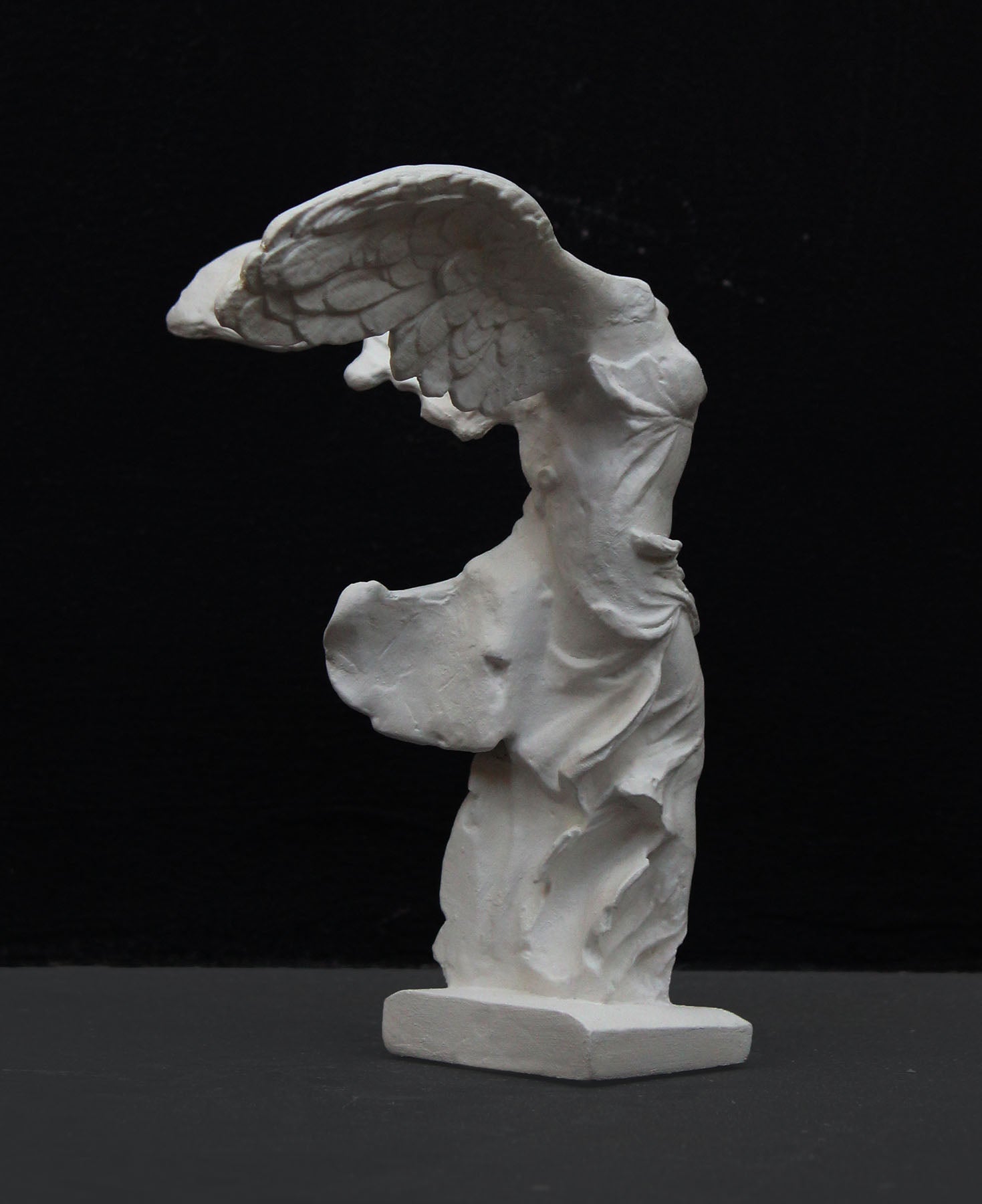photo of white plaster cast sculpture of winged, headless female figure with flowing drapery on a gray background