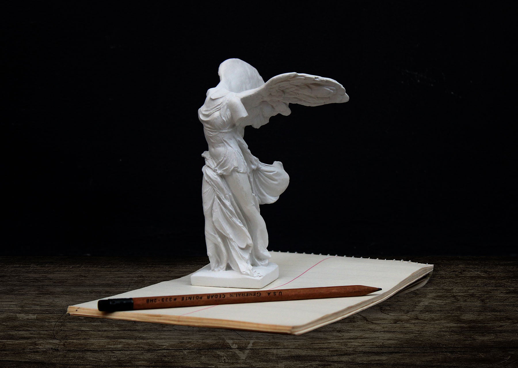 photo of white plaster cast sculpture of winged, headless female figure with flowing drapery next to a pencil atop an open notebook on a wood surface against a gray background