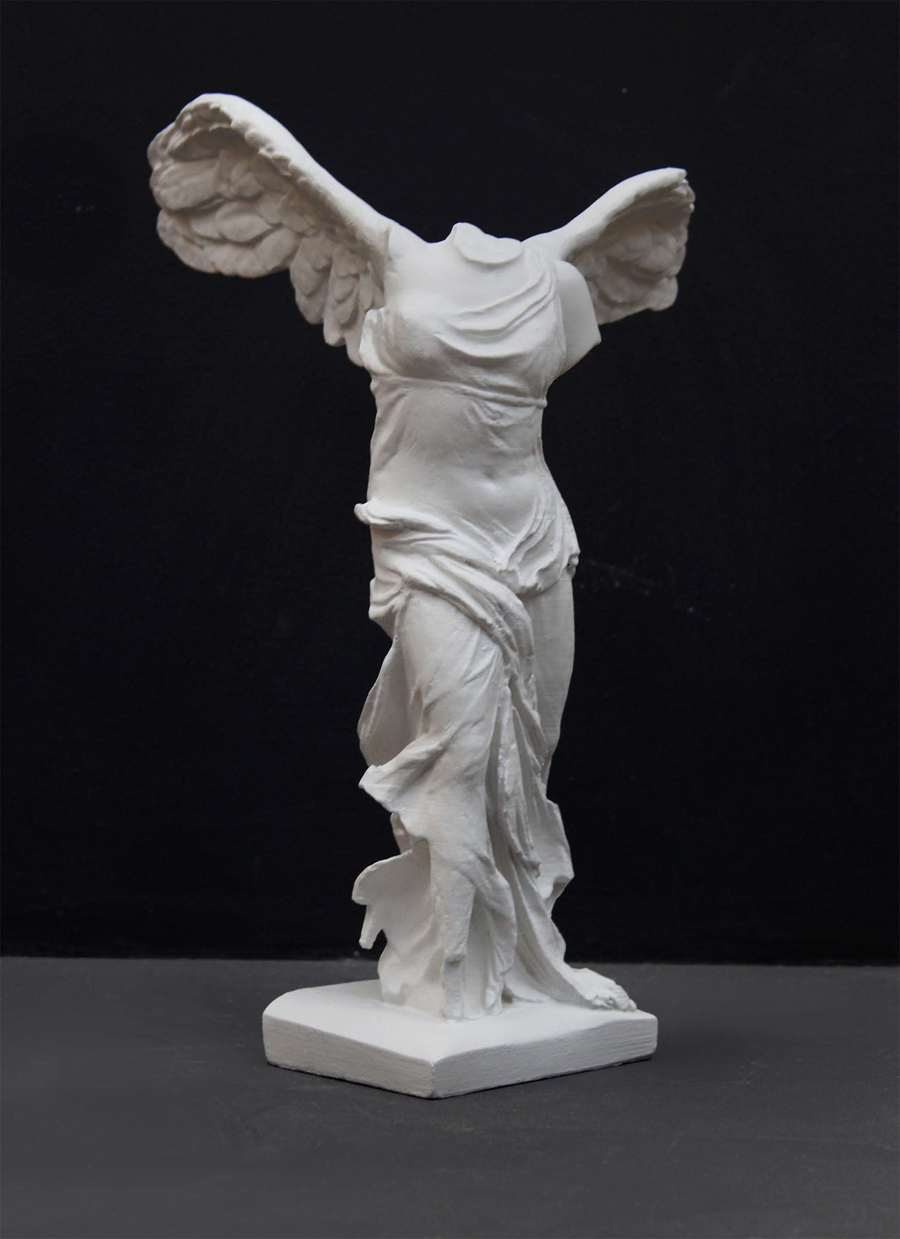 photo of white plaster cast sculpture of winged, headless female figure with flowing drapery on a gray background