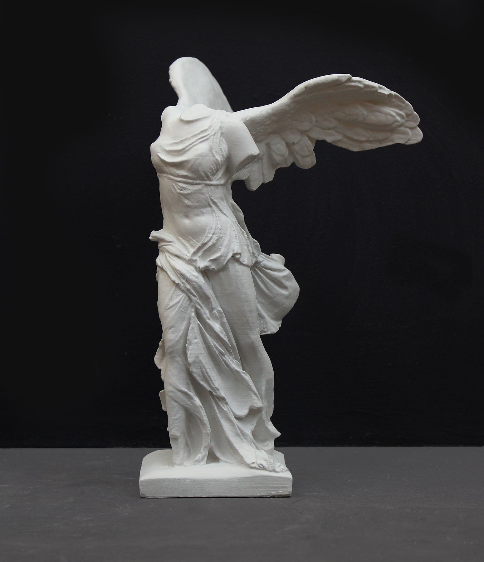 photo of white plaster cast sculpture of winged, headless female figure with flowing drapery on a gray background