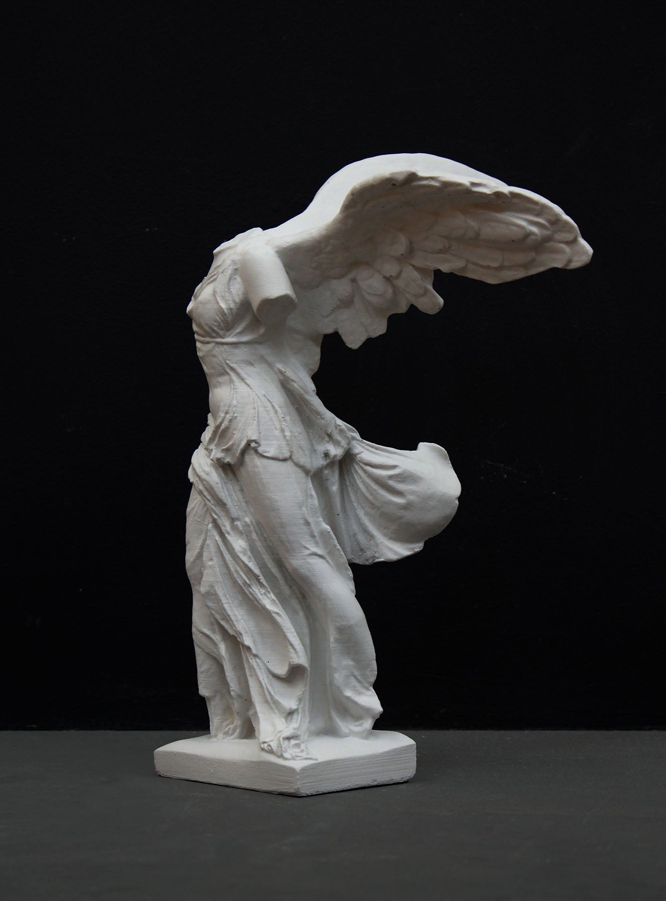 photo of white plaster cast sculpture of winged, headless female figure with flowing drapery on a gray background