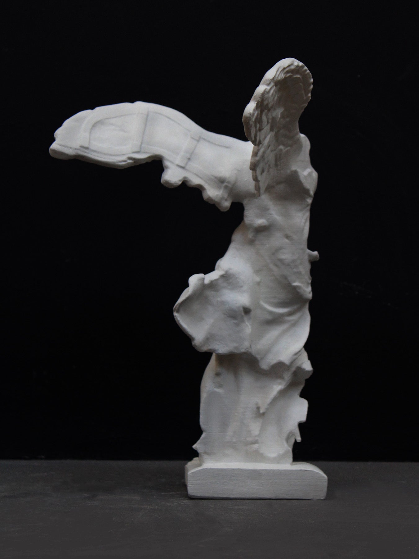 back view photo of white plaster cast sculpture of winged, headless female figure with flowing drapery on a gray background