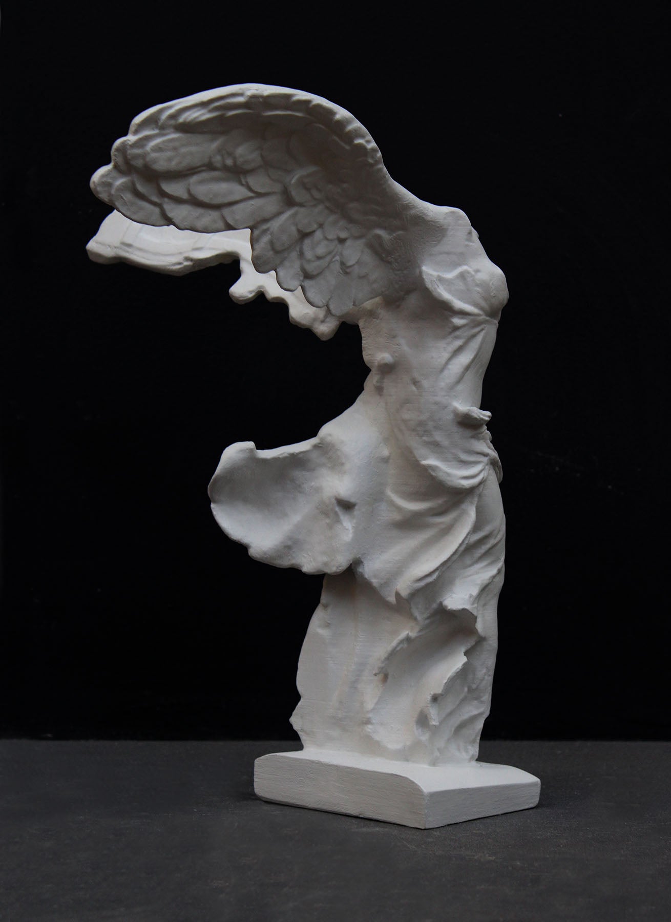 photo of white plaster cast sculpture of winged, headless female figure with flowing drapery on a gray background