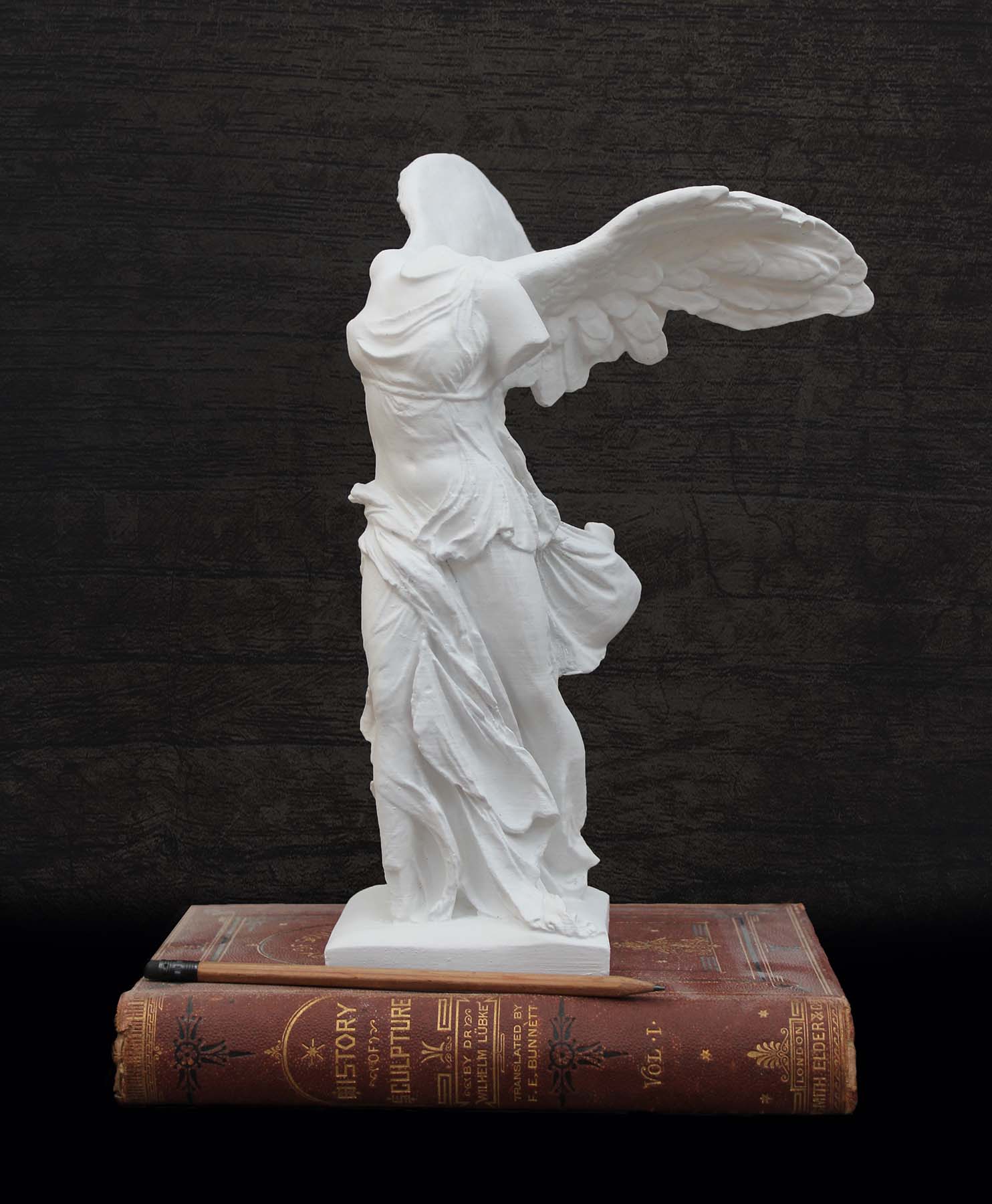 photo of white plaster cast sculpture of winged, headless female figure with flowing drapery next to a pencil atop an antique red book against a dark gray wood background