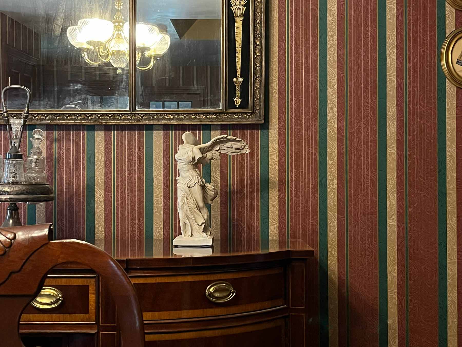photo of off-white plaster cast sculpture of winged, headless female figure with flowing drapery atop brown antique buffet with wall mirror behind and striped wallpaper behind