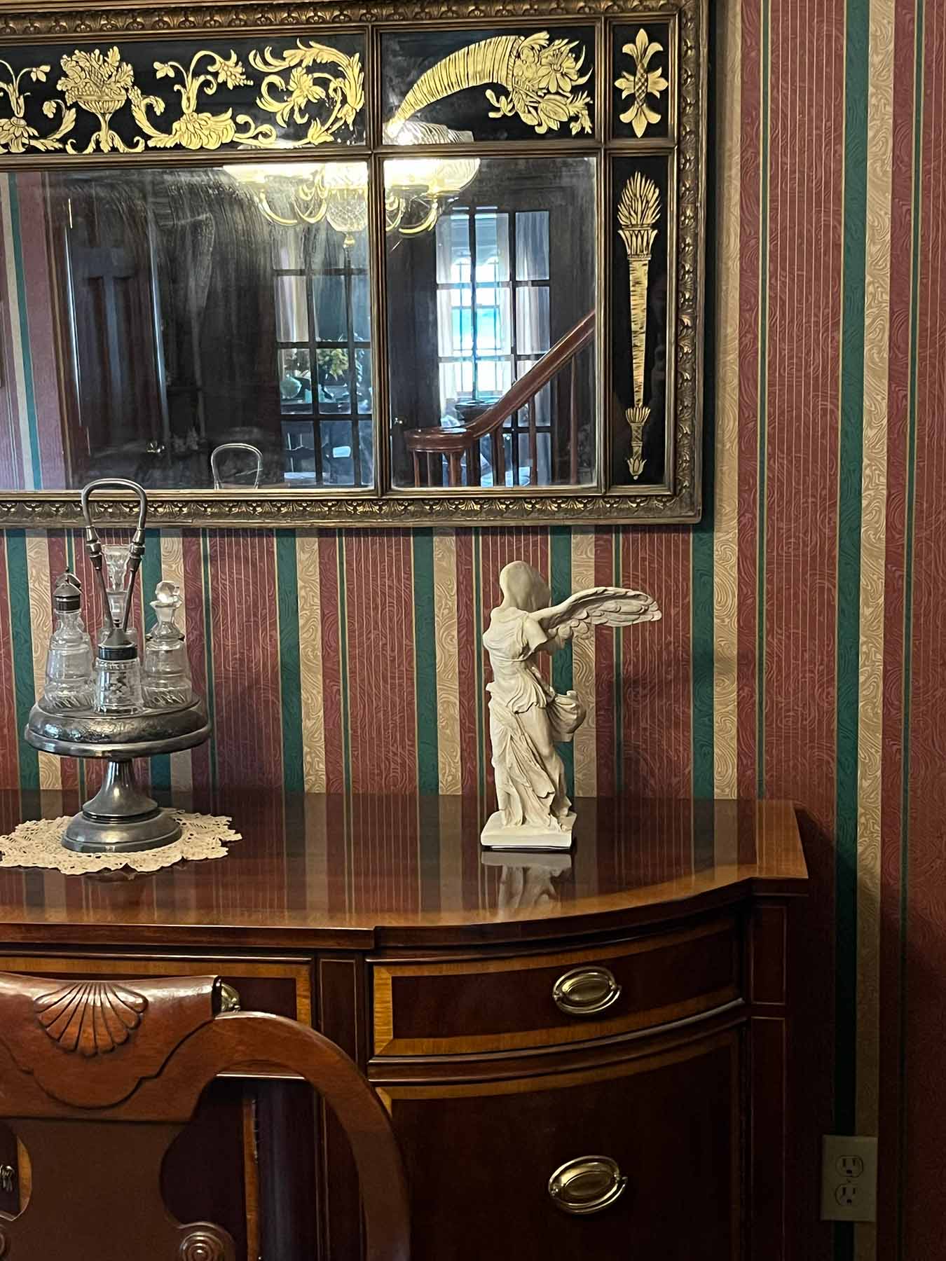 photo of off-white plaster cast sculpture of winged, headless female figure with flowing drapery atop brown antique buffet with wall mirror behind and striped wallpaper behind