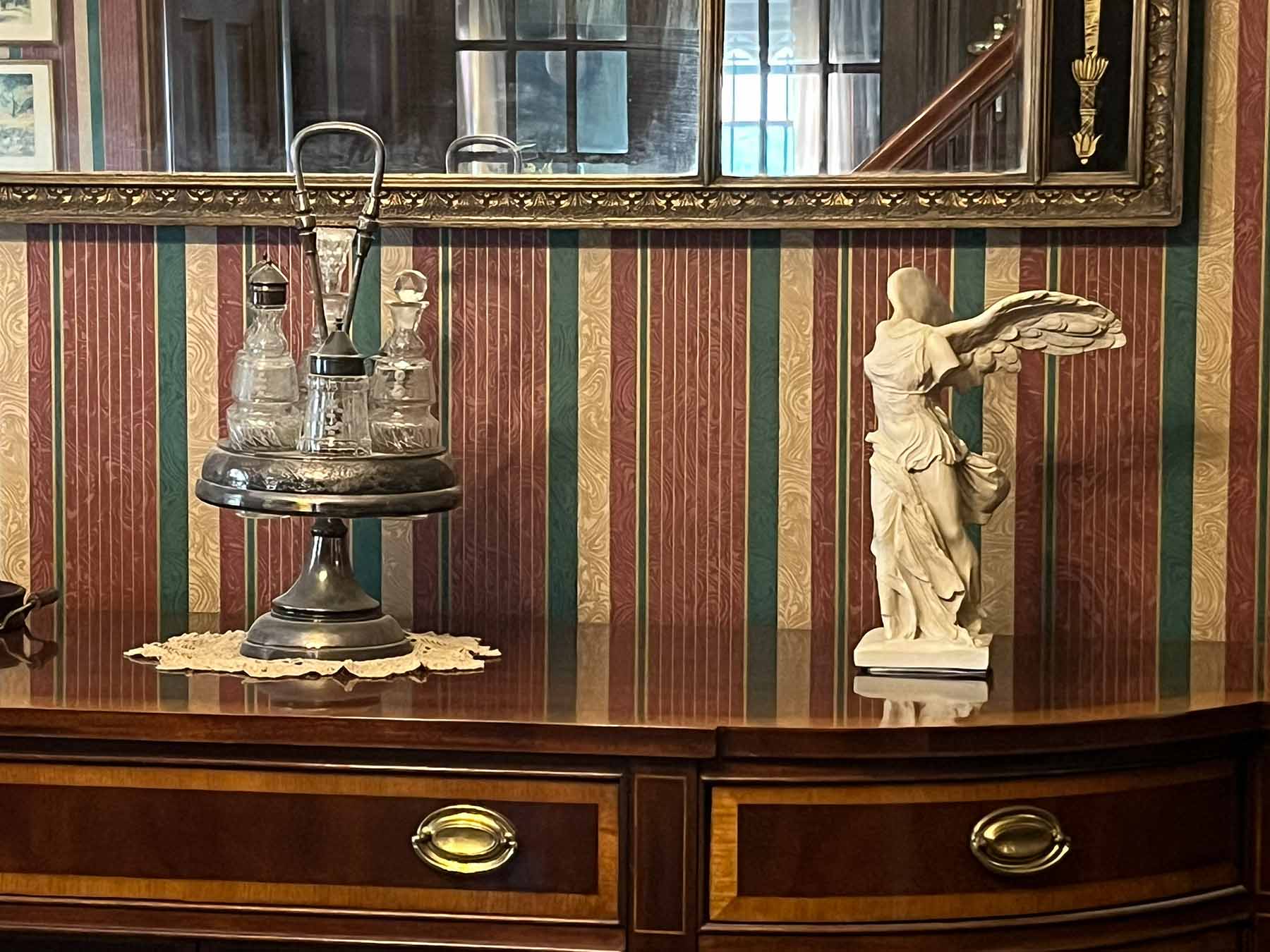 photo of off-white plaster cast sculpture of winged, headless female figure with flowing drapery atop brown antique buffet with wall mirror behind and striped wallpaper behind