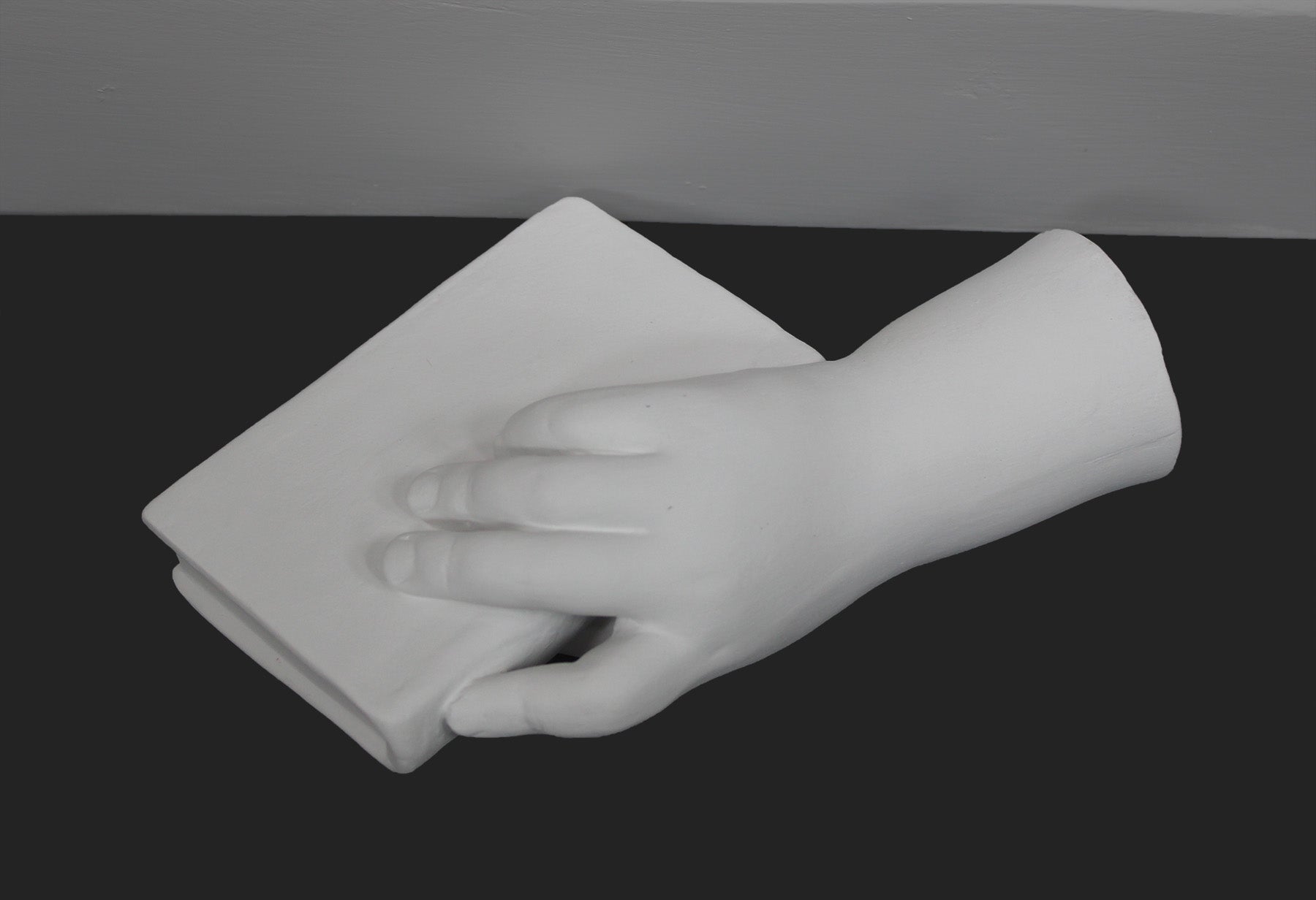 photo of white plaster cast of baby hand holding a book by the spine against gray background