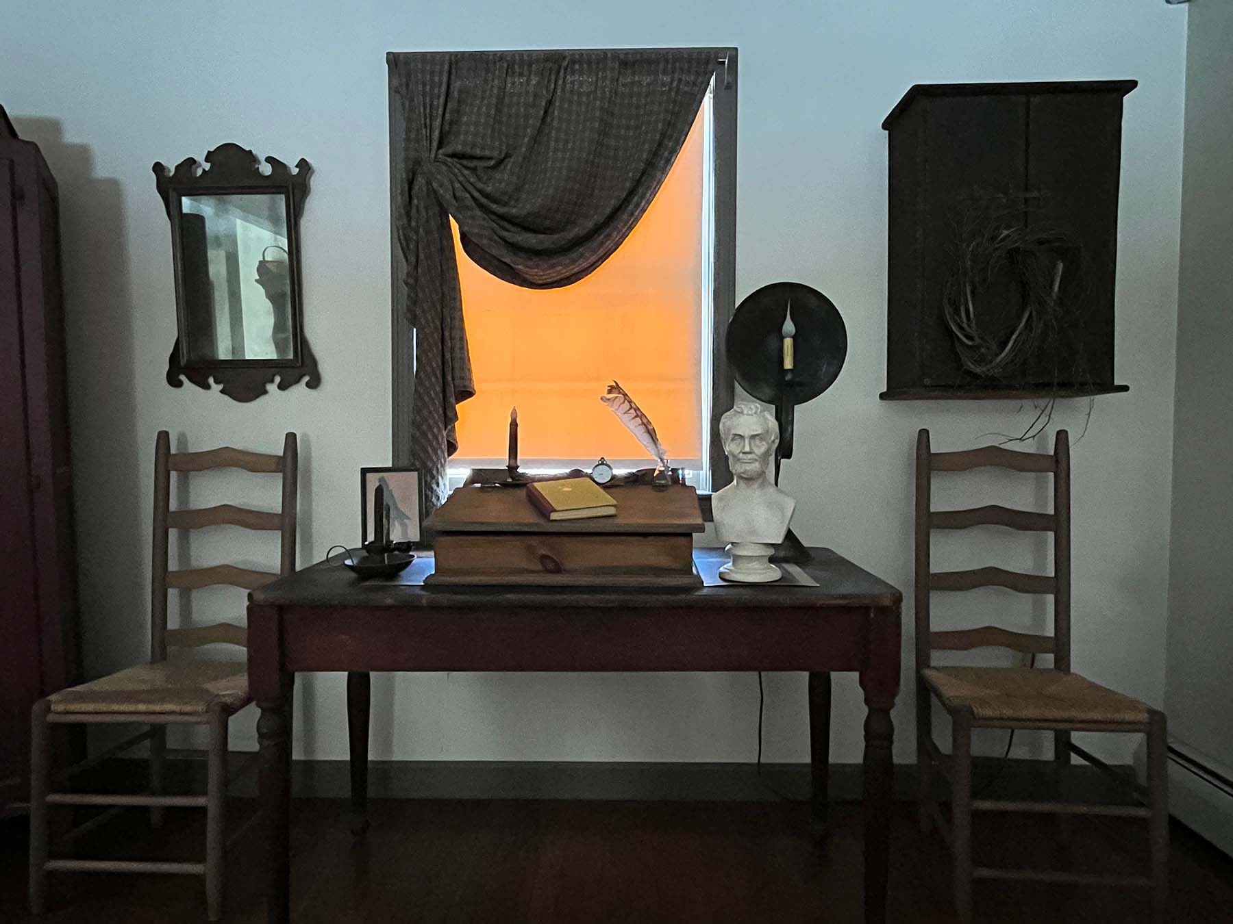 plaster cast bust sculpture of man with beard, namely Lincoln, on antique wooden table decorated with antique books, candles, and other paraphernalia and two wood and wicker chairs beside the table and sunlight coming in shaded window behind