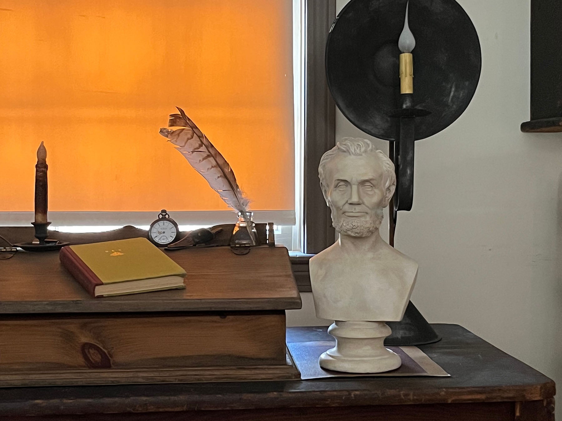 plaster cast bust sculpture of man with beard, namely Lincoln, on antique wooden table decorated with antique books, candles, and other paraphernalia and two wood and wicker chairs beside the table and sunlight coming in shaded window behind