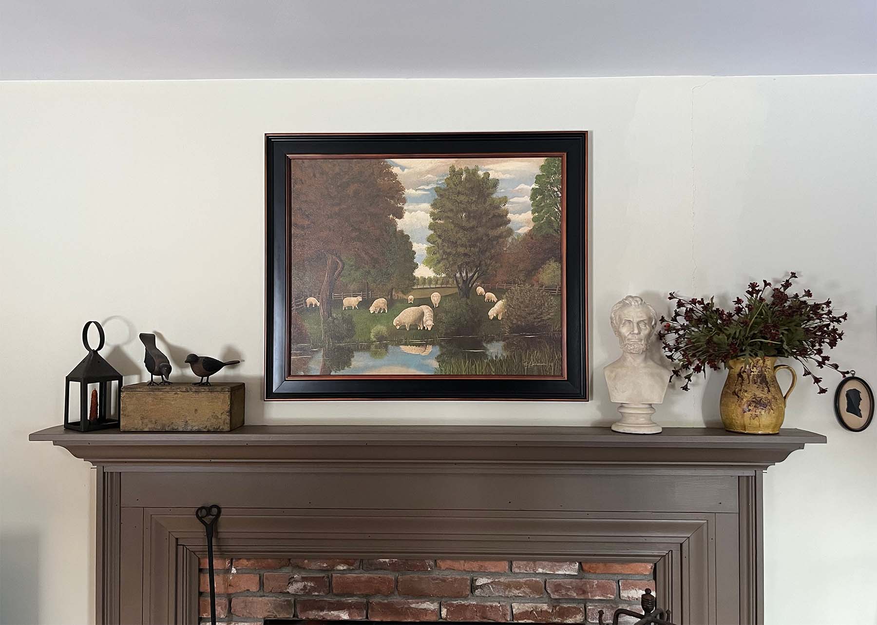 plaster cast bust sculpture of man with beard, namely Lincoln, on mantel beside cream-colored jar holding small red flowers and folk art painting of animals on white wall behind