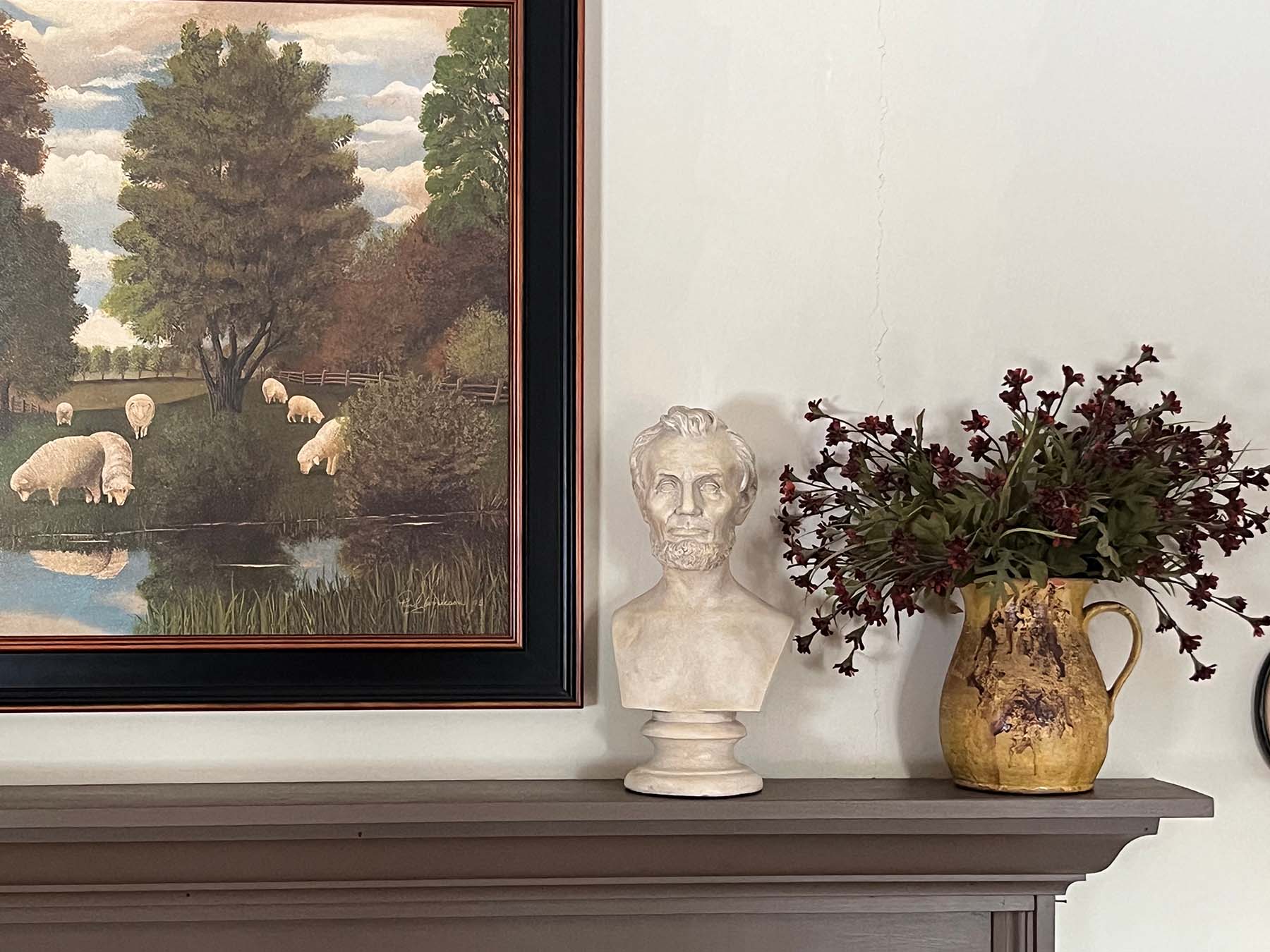 plaster cast bust sculpture of man with beard, namely Lincoln, on mantel beside cream-colored jar holding small red flowers and folk art painting of animals on white wall behind