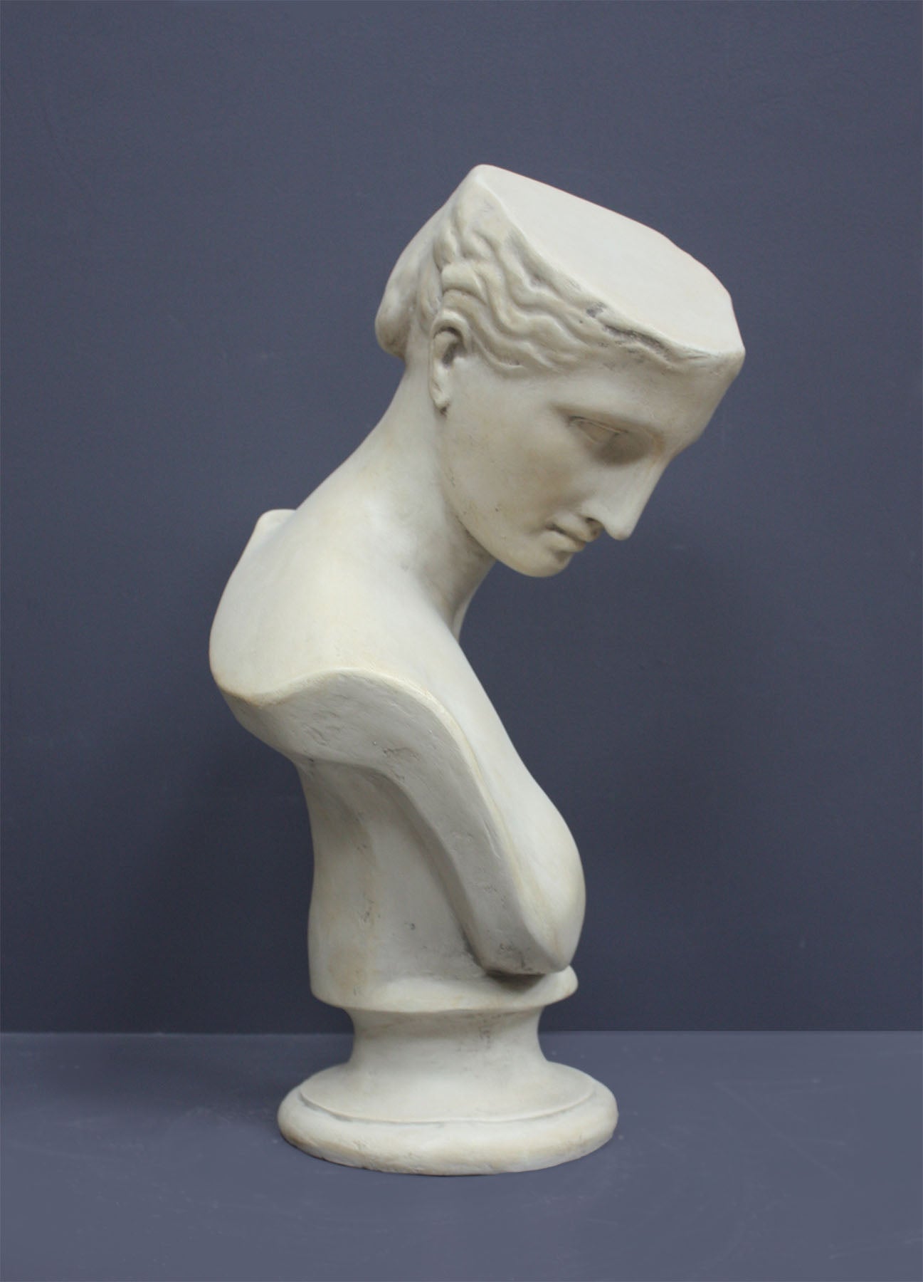 photo of off-white plaster cast sculpture bust of female without top of head and with piece of drapery on socle base on gray background
