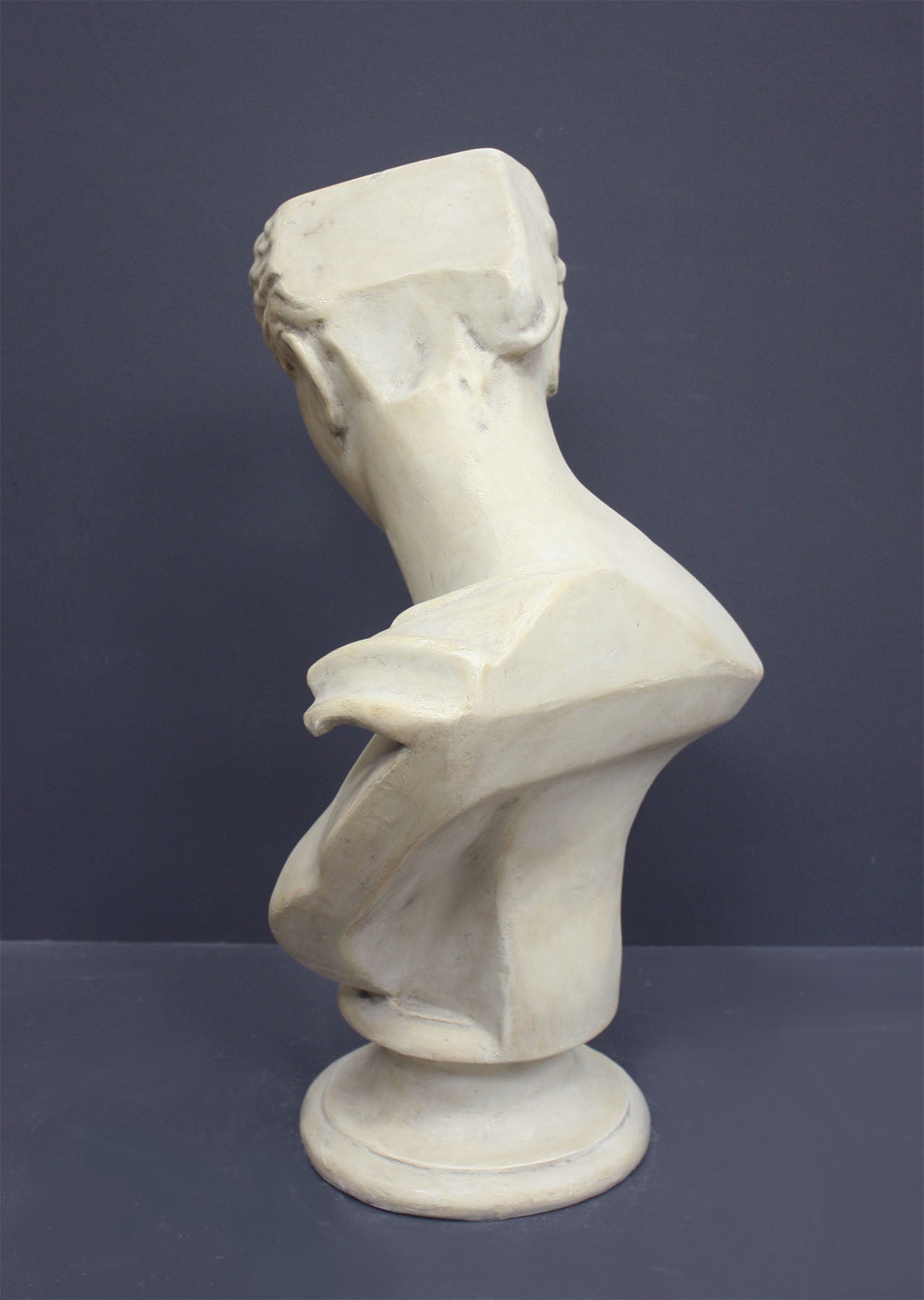 photo of off-white plaster cast sculpture bust of female without top of head and with piece of drapery on socle base on gray background