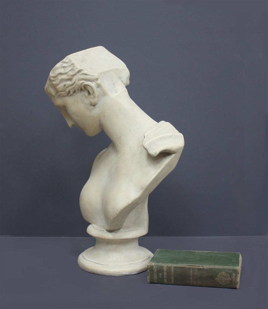 photo of off-white plaster cast sculpture bust of female without top of head and with piece of drapery on socle base with green book laying beside it on gray background