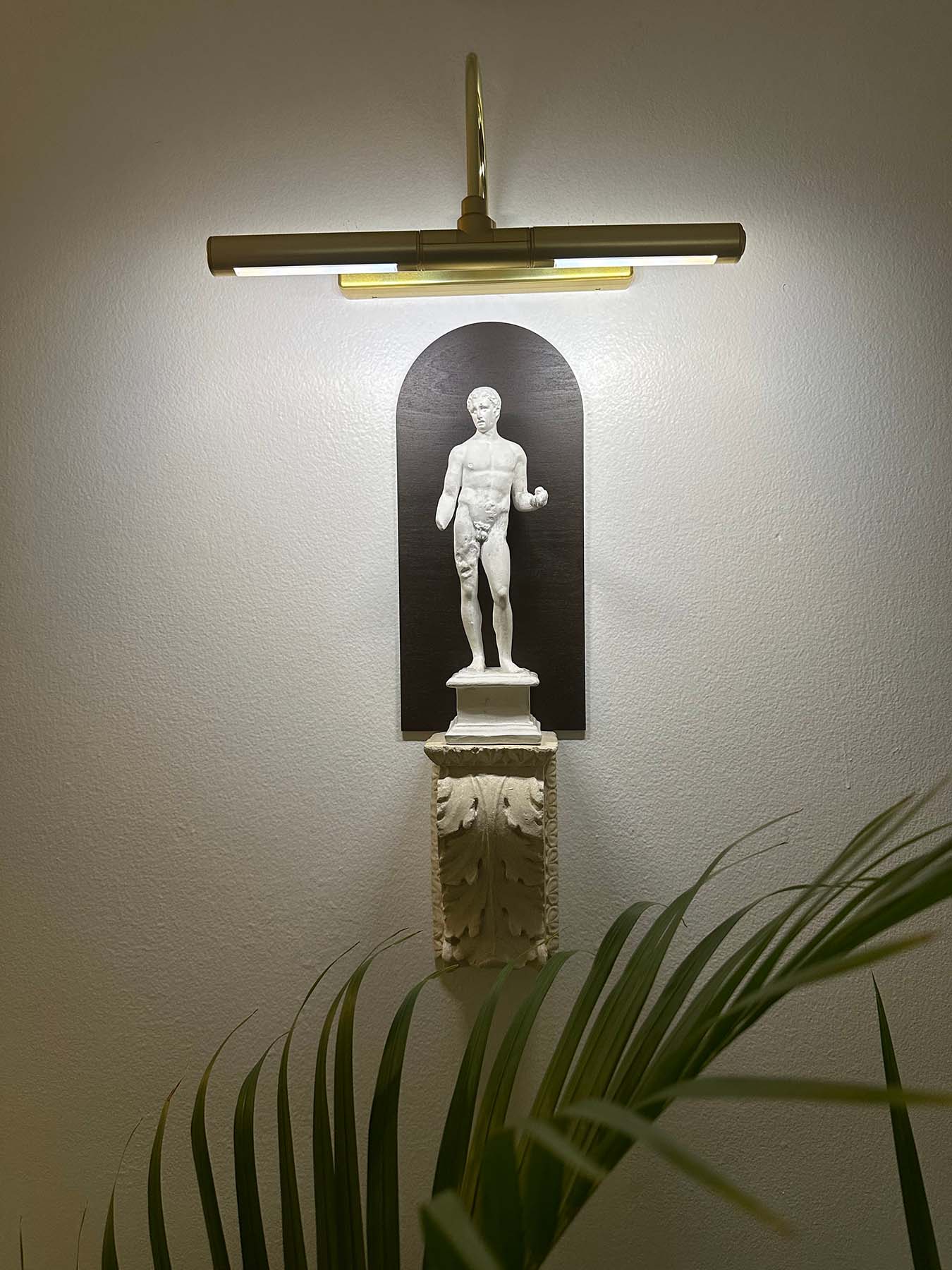 photo of plaster cast sculpture of classical male figure, namely Hercules, wall-mounted atop a plaster cast bracket with leaf design and top lit for nighttime viewing on a neutral wall and plant leaves visible in foreground