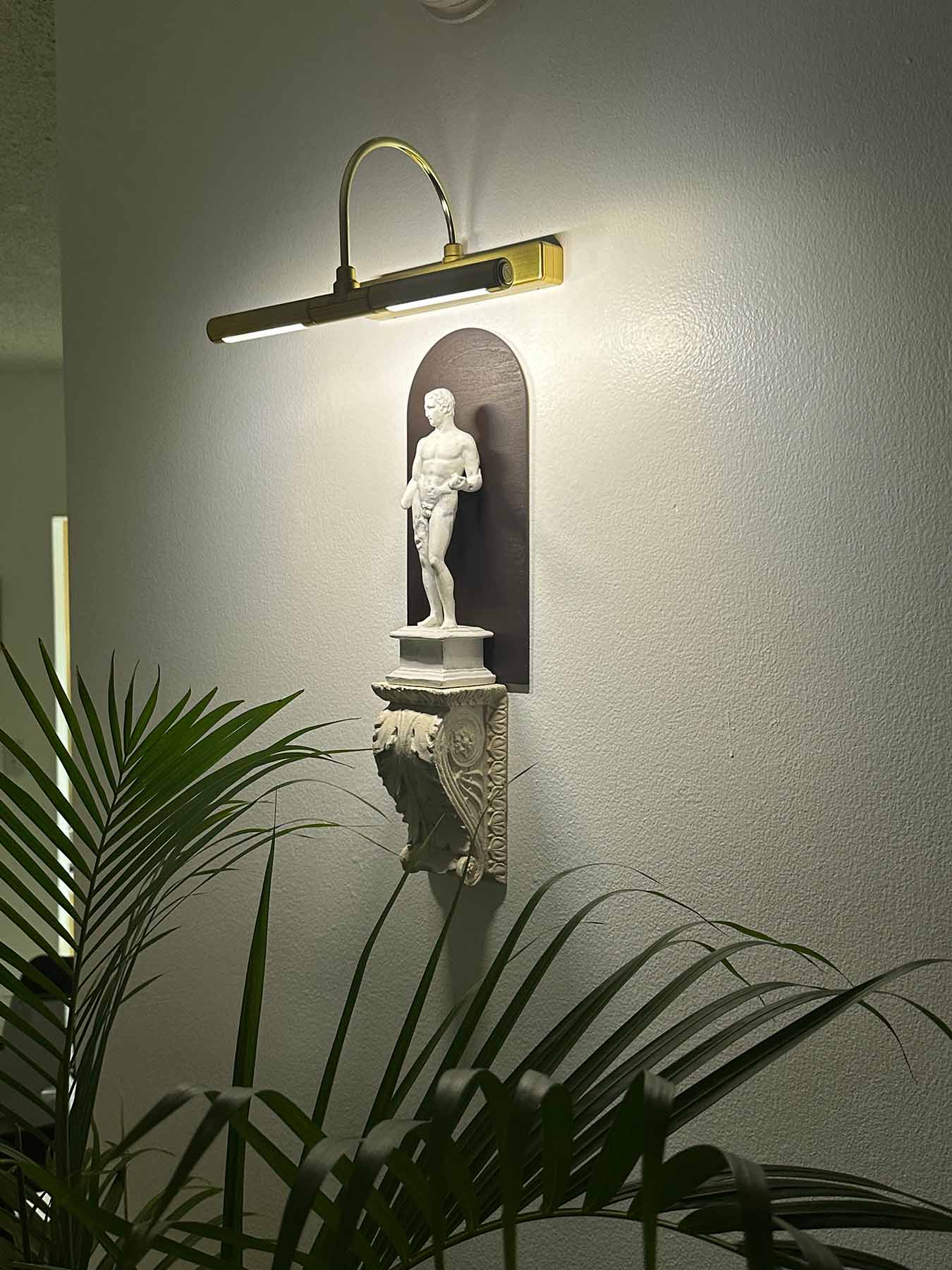 photo of plaster cast sculpture of classical male figure, namely Hercules, wall-mounted atop a plaster cast bracket with leaf design and top lit for nighttime viewing on a neutral wall and plant leaves visible in foreground