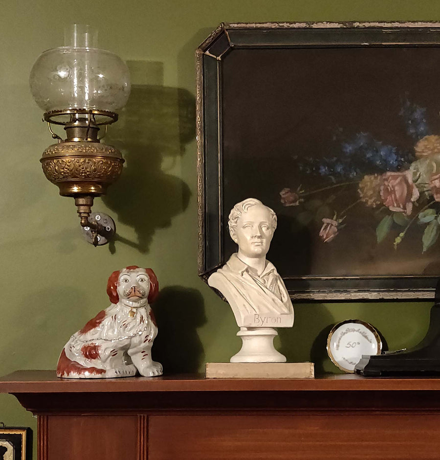 photo of off-white plaster cast sculpture bust of man, namely the poet Lord Byron, on socle base atop book on mantel beside foo dog with painting and antique globe light on wall