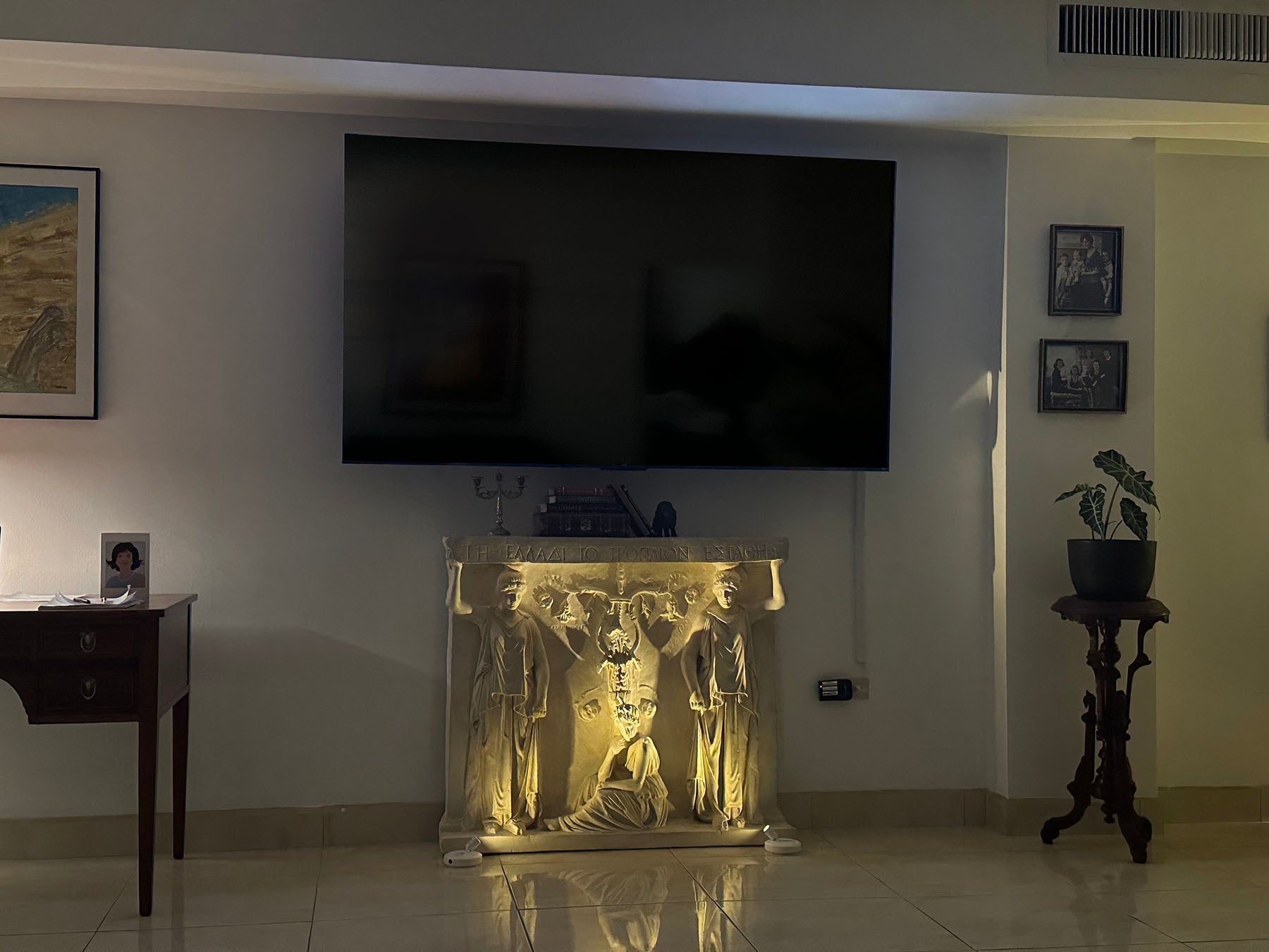 photo of plaster cast of free-standing relief sculpture with three female figures, two of which are caryatides, against a living room wall with a television mounted above it and wooden desk and other decor nearby, and relief floor lit for nighttime viewing