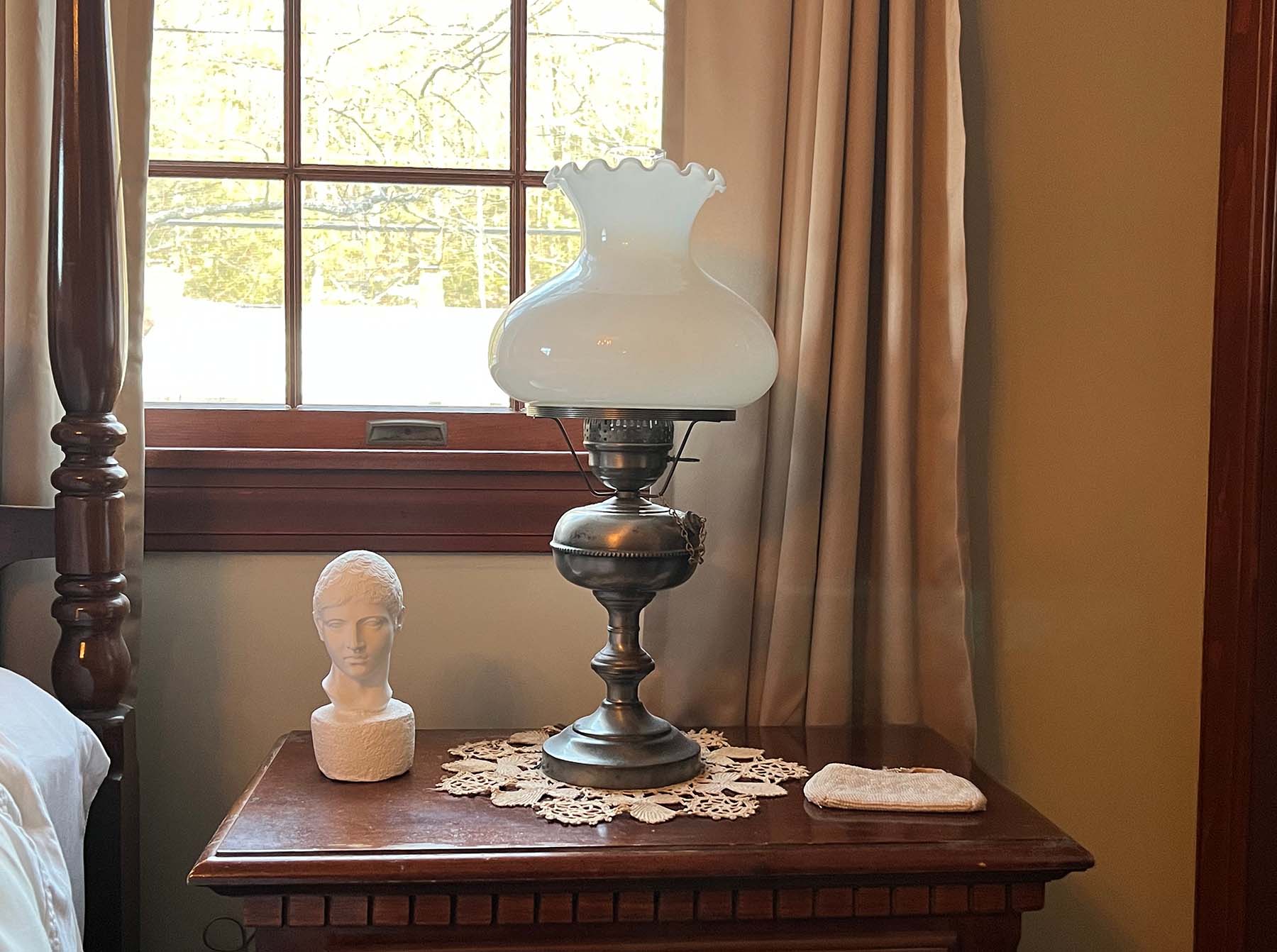 photo of white plaster cast head of male on brown nightstand beside silver lamp with milk glass in front of window