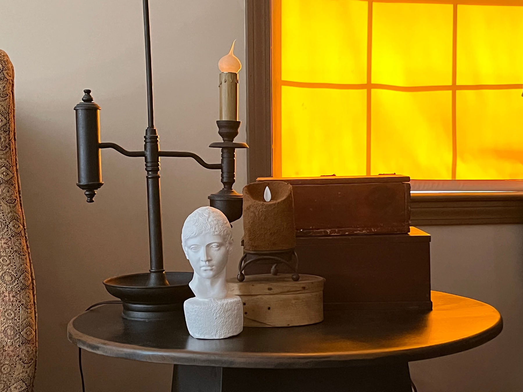 photo of white plaster cast head of male on small round table decorated with wood boxes and pillar candle and candelabra with sunlight coming in shaded window behind