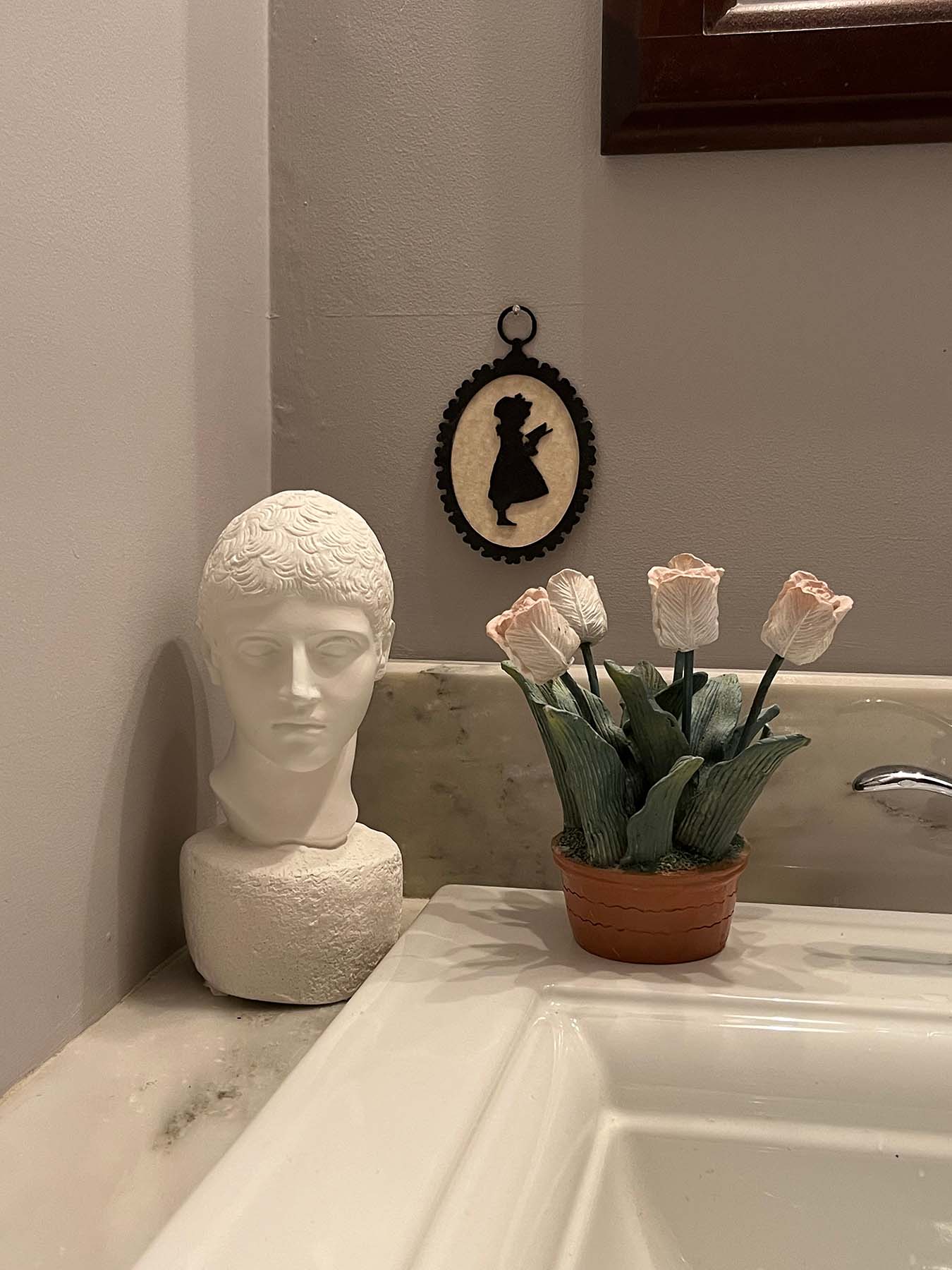 photo of white plaster cast head of male on white marble sink counter with white sink and ceramic flowers beside it and small silhouette of girl behind on gray wall