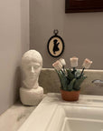 photo of white plaster cast head of male on white marble sink counter with white sink and ceramic flowers beside it and small silhouette of girl behind on gray wall
