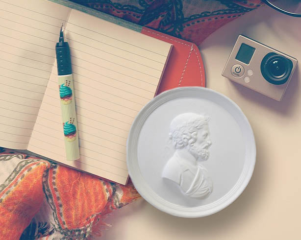 Photo of a white plaster cast of a medallion with a profile of a man wearing a wrath on his head, on a surface next to an open blank diary, pen, camera and orange patterned scarf.