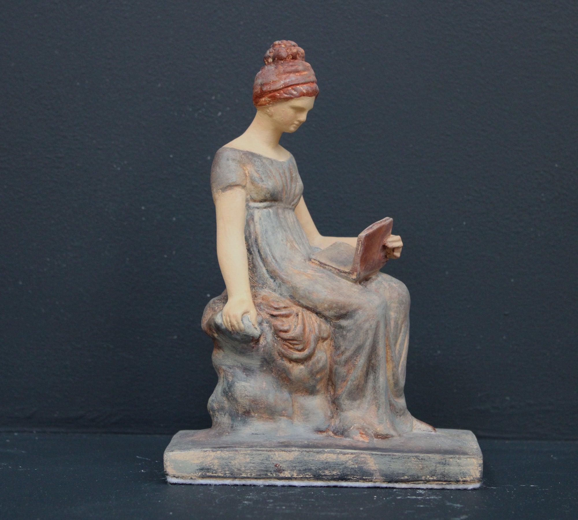 Photo of plaster cast sculpture of female in green-brown tinted robes seated with opened book against grey background