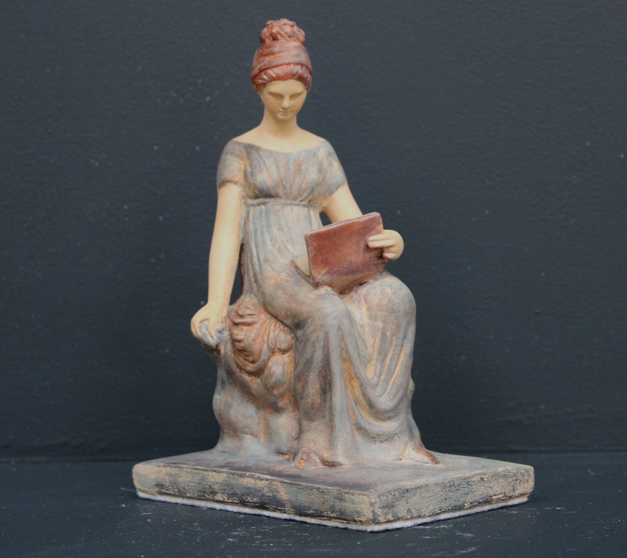 Photo of plaster cast sculpture of female in green-brown tinted robes seated with opened book against grey background