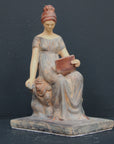 Photo of plaster cast sculpture of female in green-brown tinted robes seated with opened book against grey background