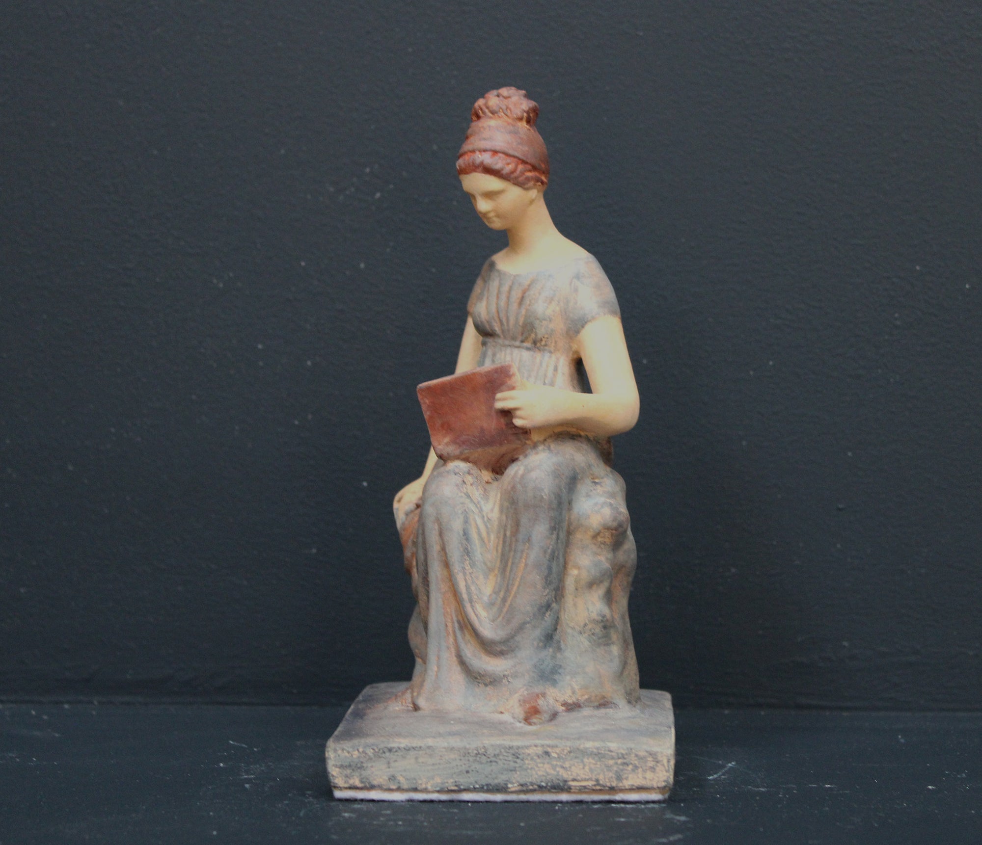 Photo of plaster cast sculpture of female in green-brown tinted robes seated with opened book against grey background
