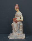 Photo of plaster cast sculpture of female in green-brown tinted robes seated with opened book against grey background