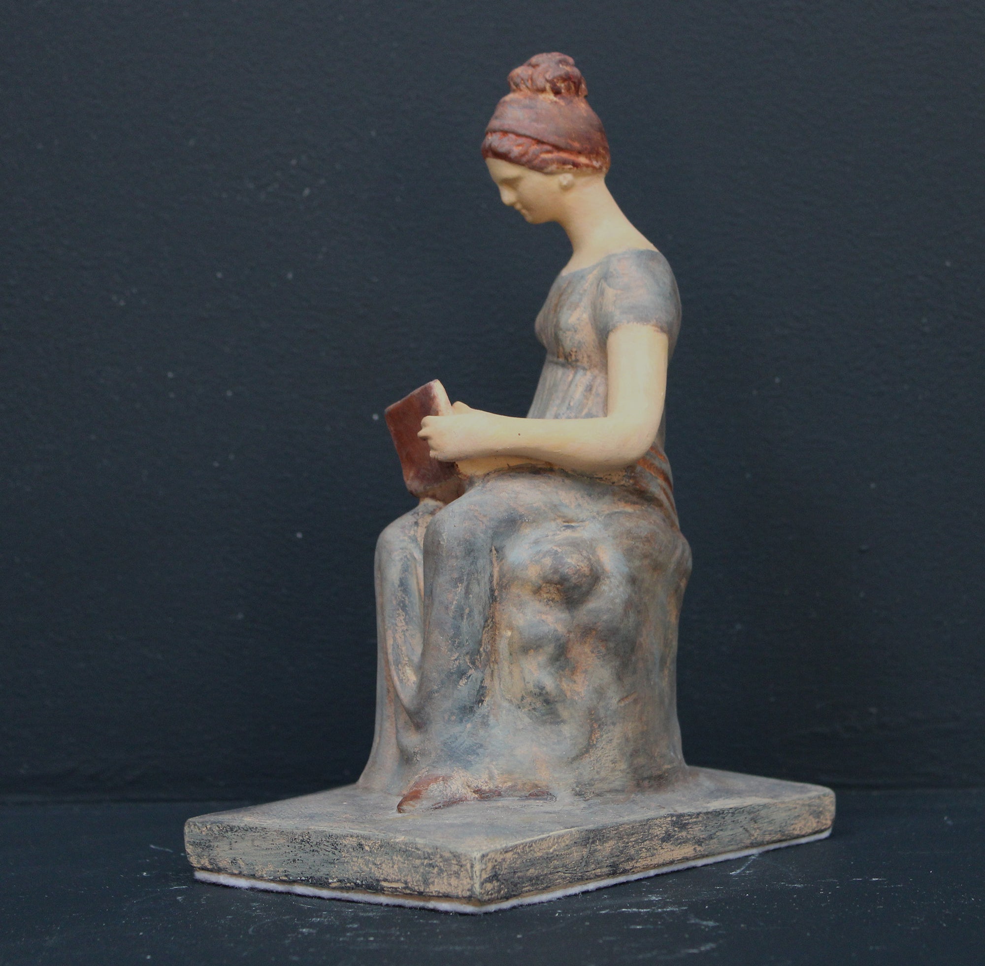 Photo of plaster cast sculpture of female in green-brown tinted robes seated with opened book against grey background