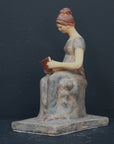 Photo of plaster cast sculpture of female in green-brown tinted robes seated with opened book against grey background