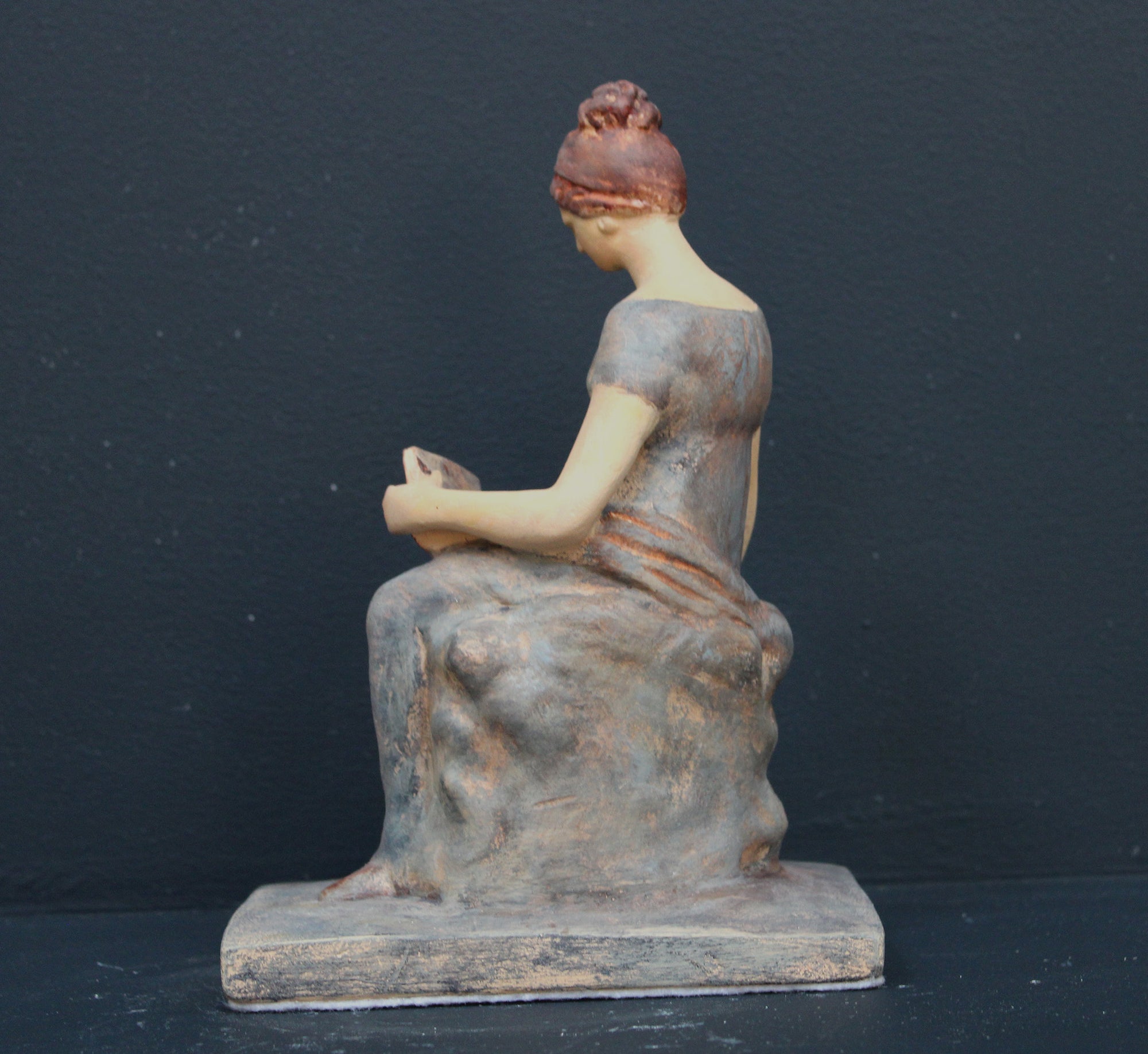 Photo of plaster cast sculpture of female in green-brown tinted robes seated with opened book against grey background