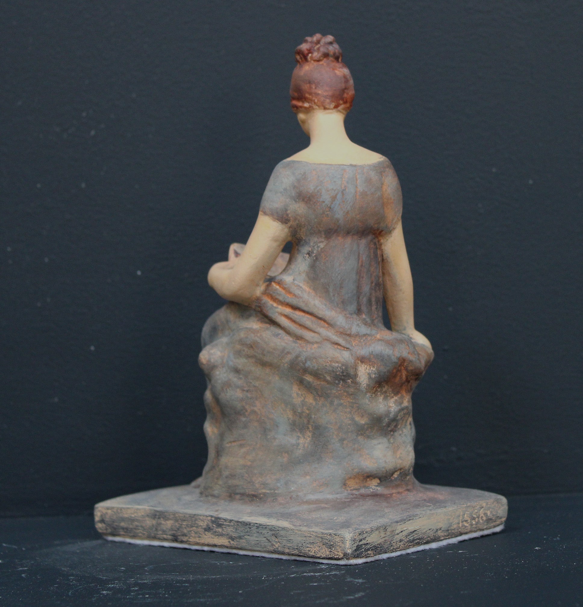 Photo of plaster cast sculpture of female in green-brown tinted robes seated with opened book against grey background