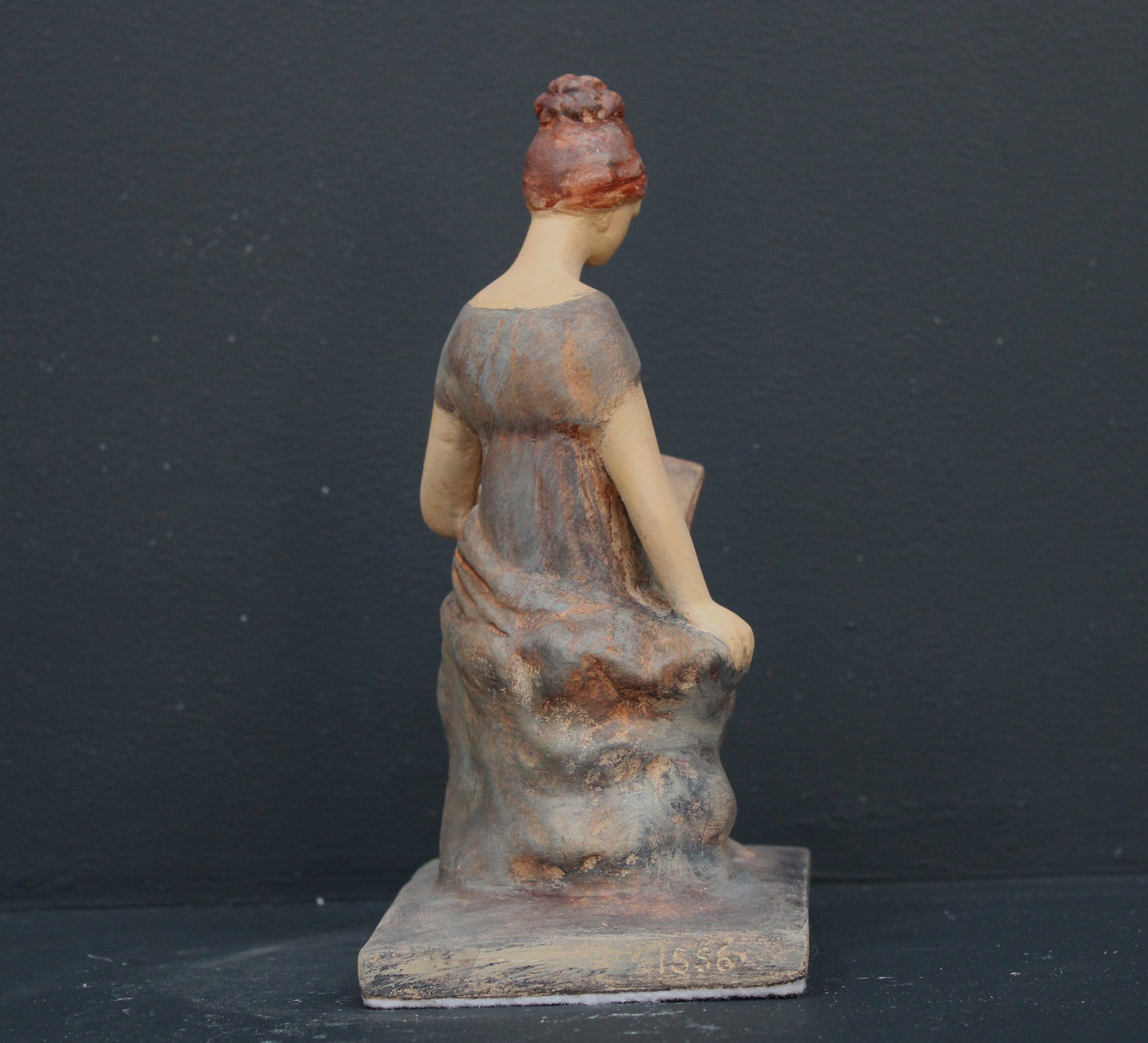 Photo of plaster cast sculpture of female in green-brown tinted robes seated with opened book against grey background