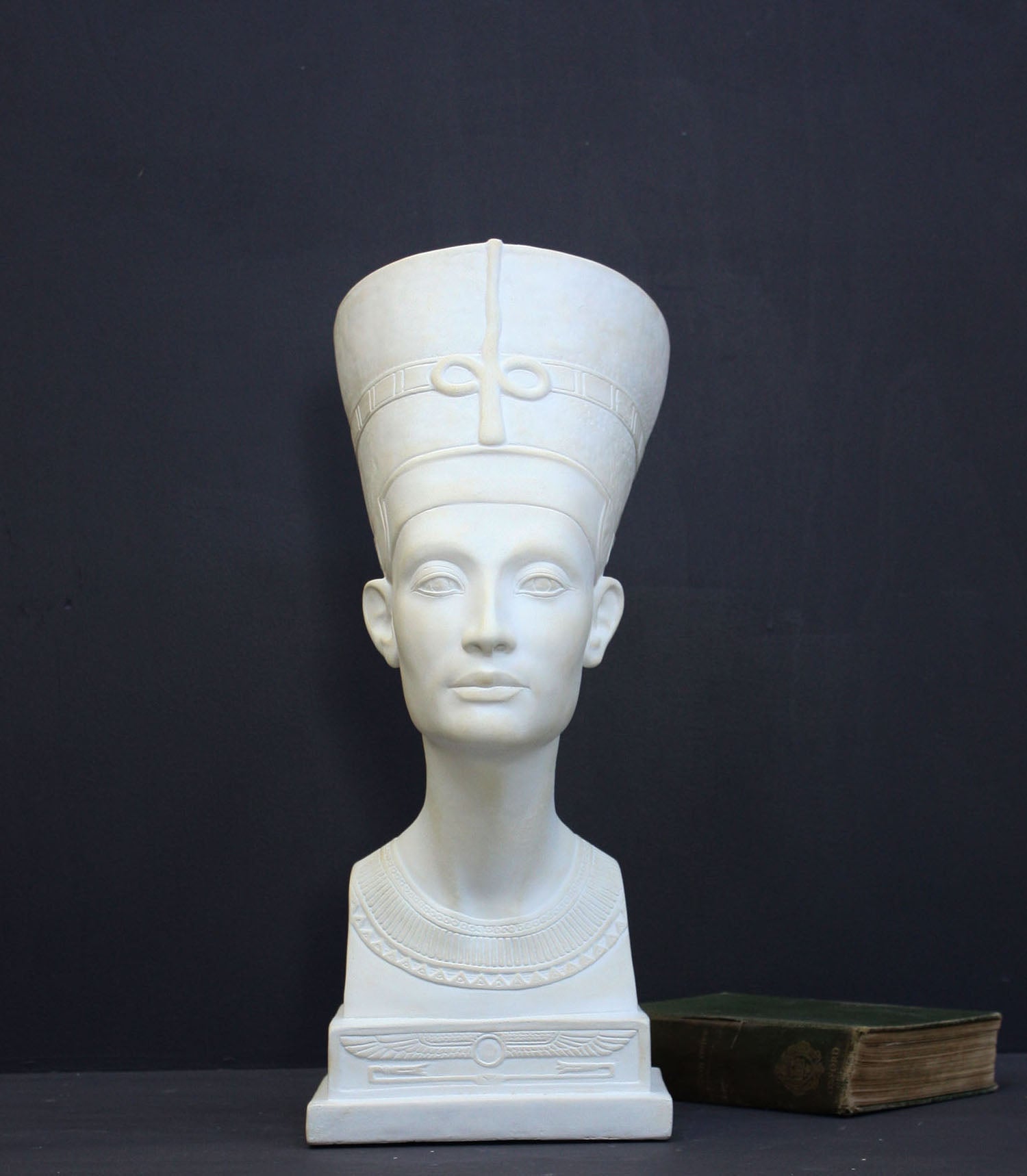 photo of plaster cast sculpture of bust of Nefertiti with crown and decorative collar on a decorative base with a black background
