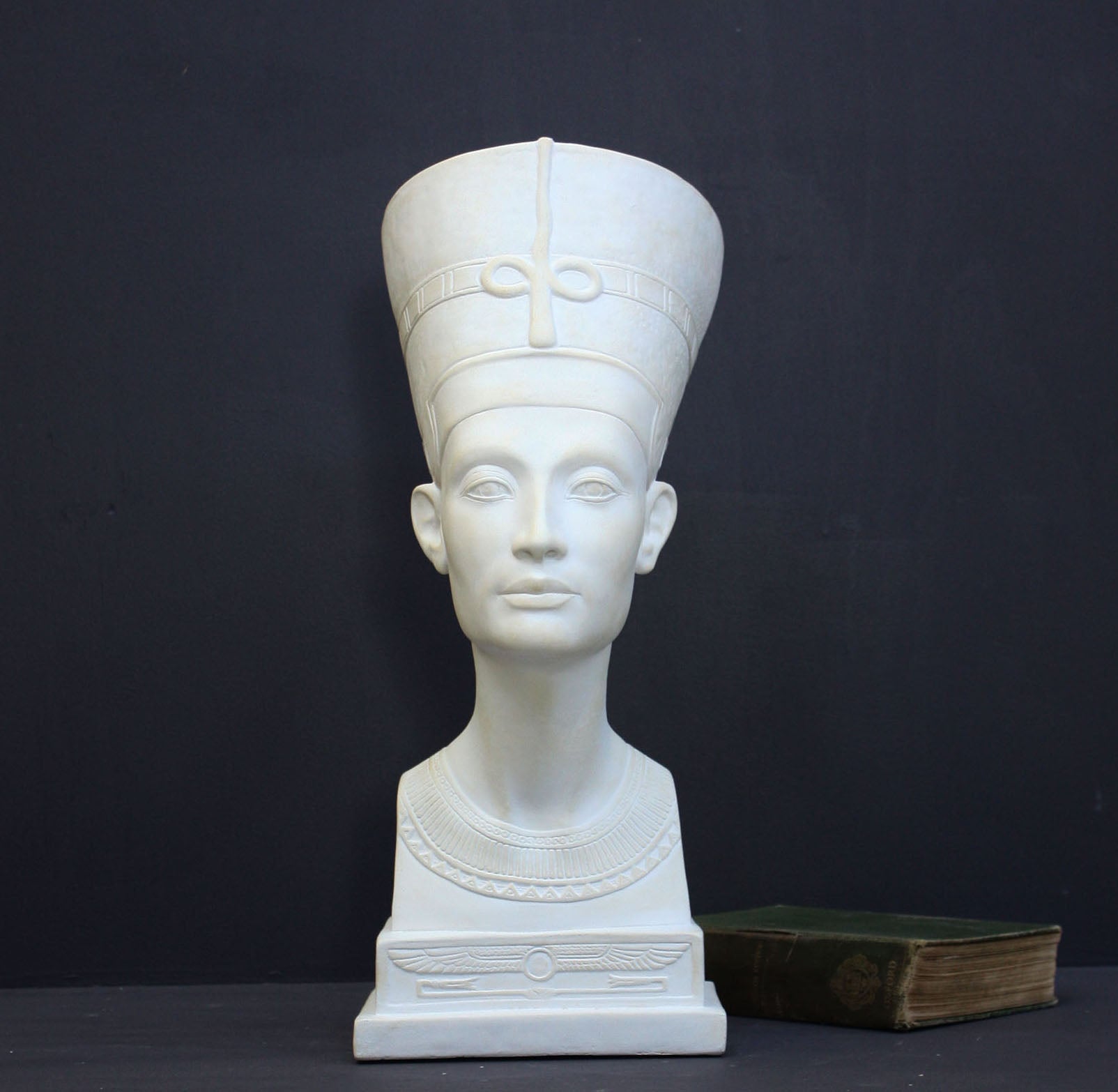 photo of plaster cast sculpture of bust of Nefertiti with crown and decorative collar on a decorative base with a black background