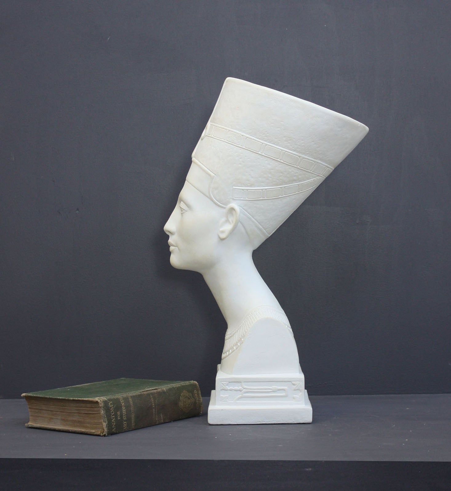 photo of plaster cast sculpture of bust of Nefertiti with crown and decorative collar on a decorative base with a black background