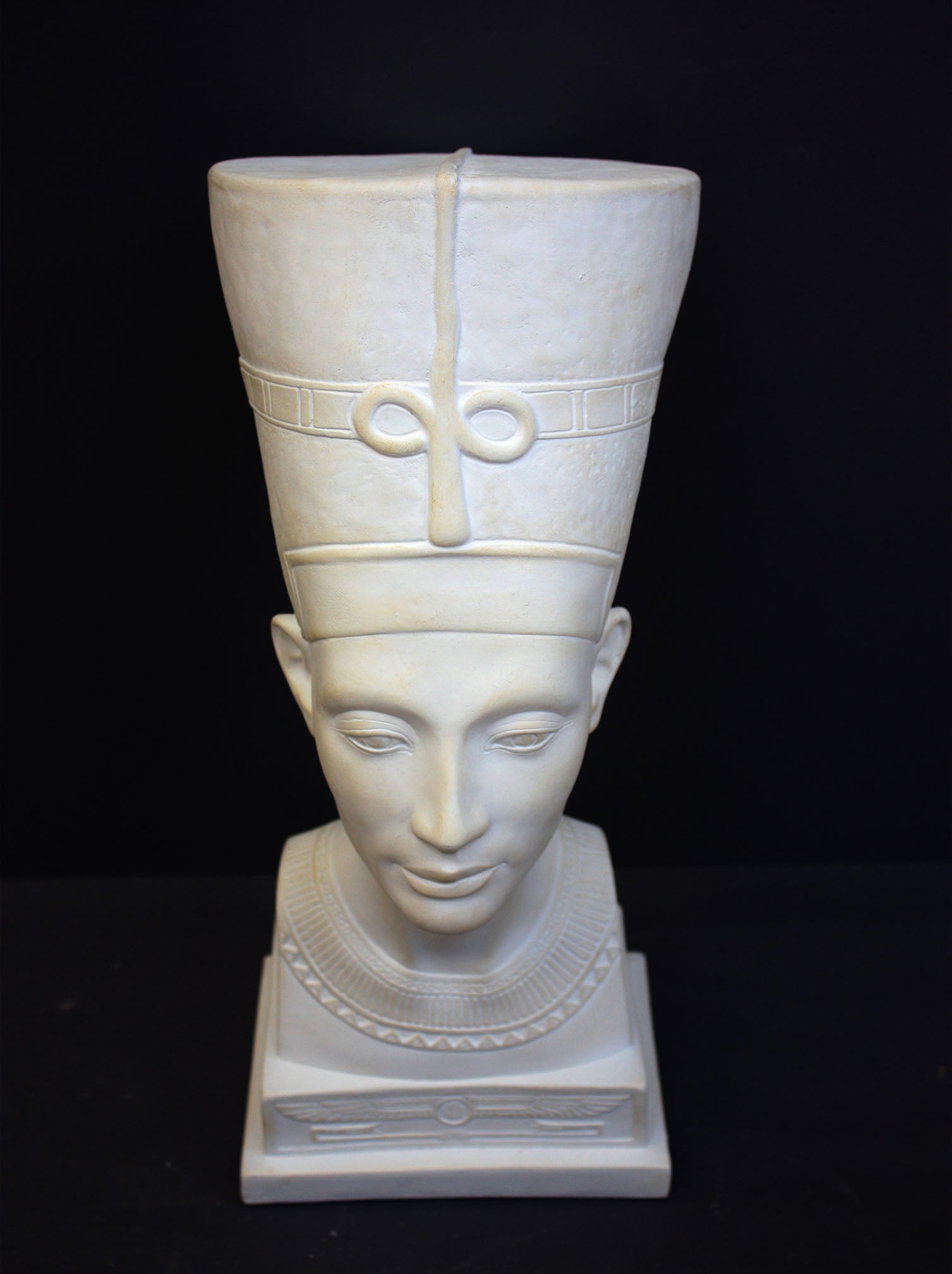 photo of plaster cast sculpture of bust of Nefertiti with crown and decorative collar on a decorative base with a black background