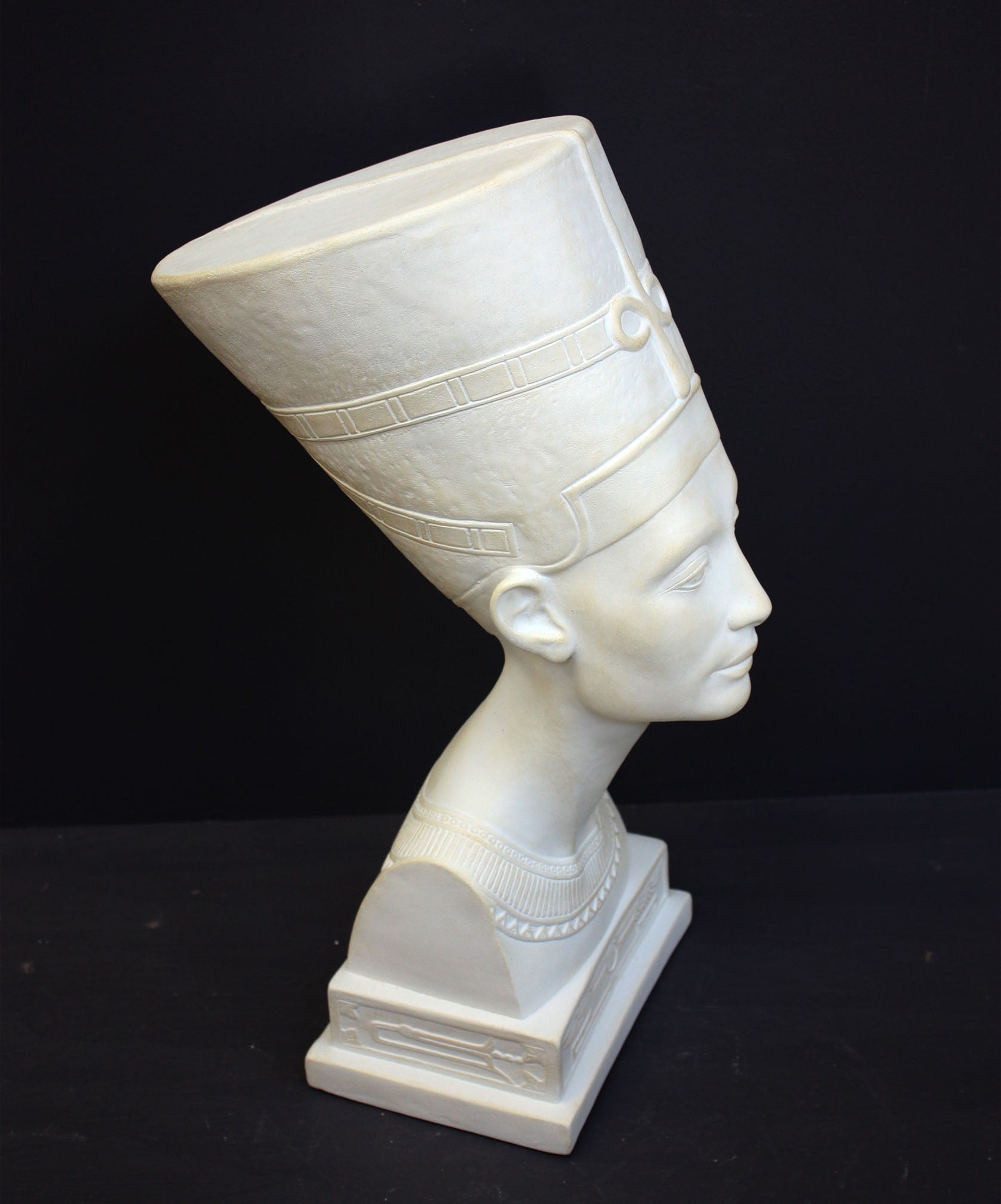 photo of plaster cast sculpture of bust of Nefertiti with crown and decorative collar on a decorative base with a black background