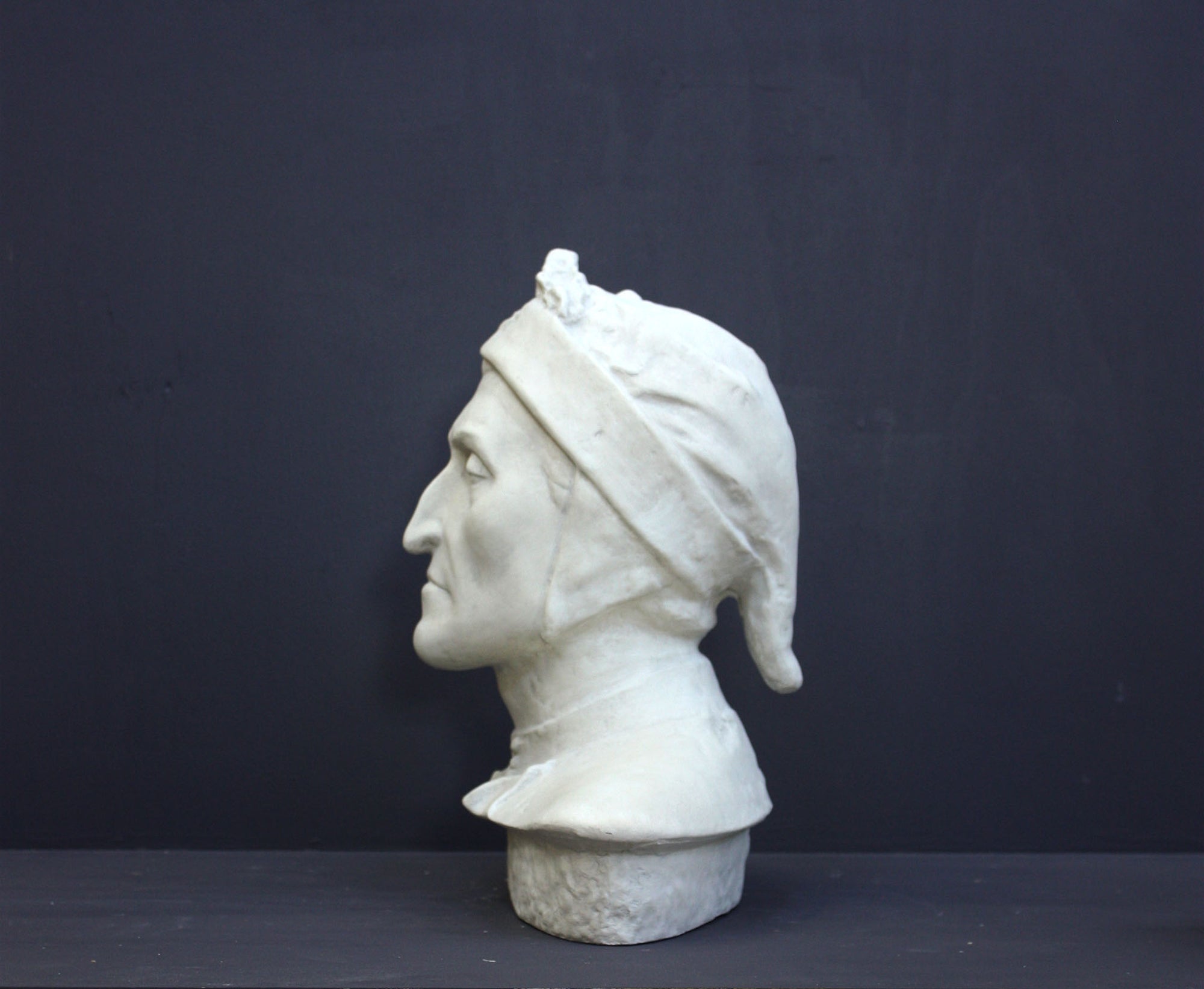 photo of plaster cast bust sculpture of Dante Alighieri with cap and high-necked robe with red book leaning against right side on black background