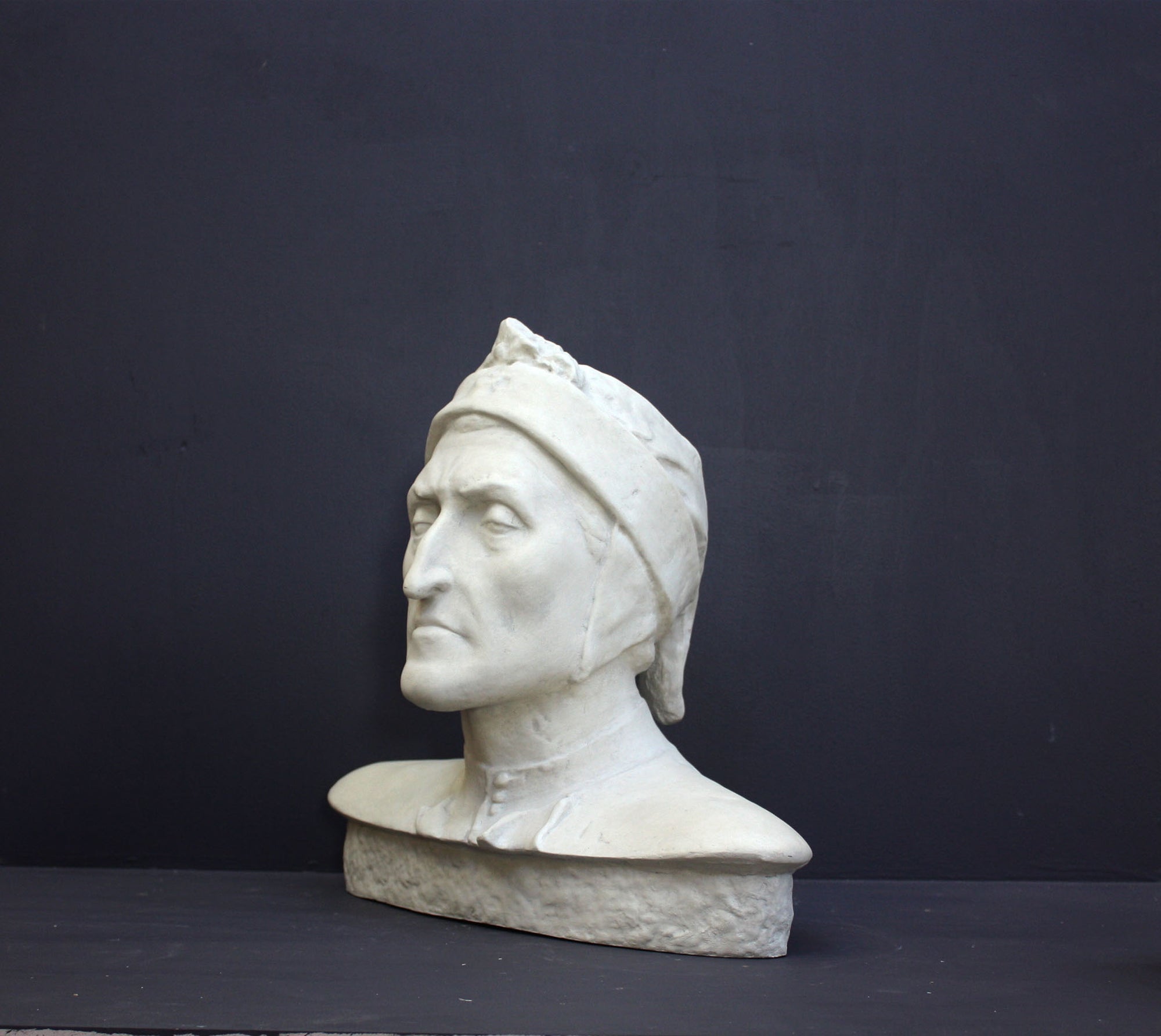 photo of plaster cast bust sculpture of Dante Alighieri with cap and high-necked robe with red book leaning against right side on black backgtround