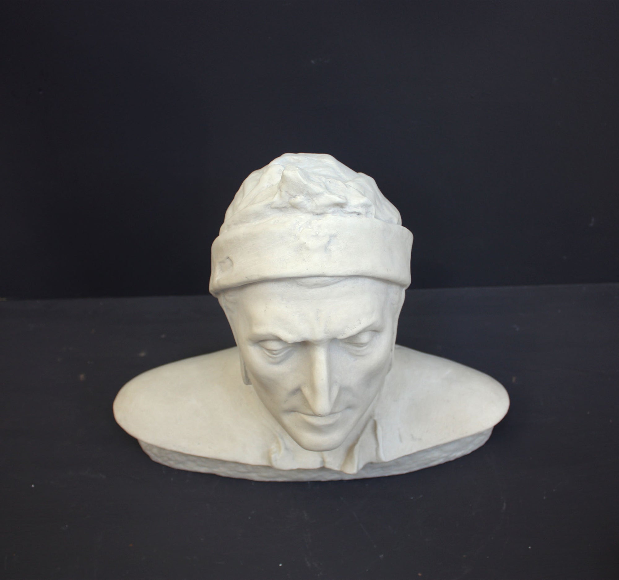 photo of plaster cast bust sculpture of Dante Alighieri with cap and high-necked robe with red book leaning against right side on black background 