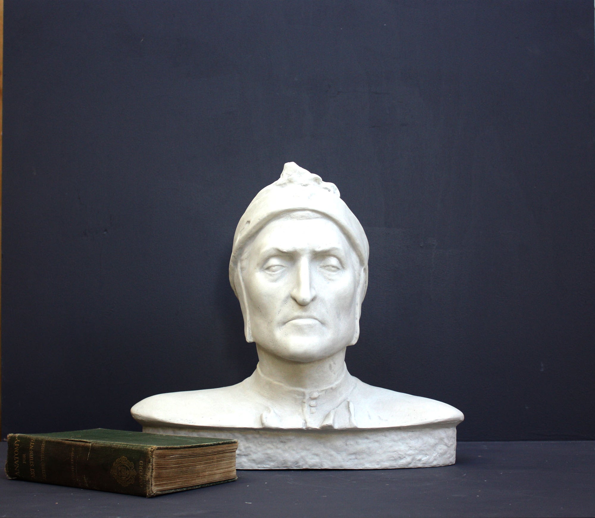 photo of plaster cast bust sculpture of Dante Alighieri with cap and high-necked robe with red book leaning against right side on black background 